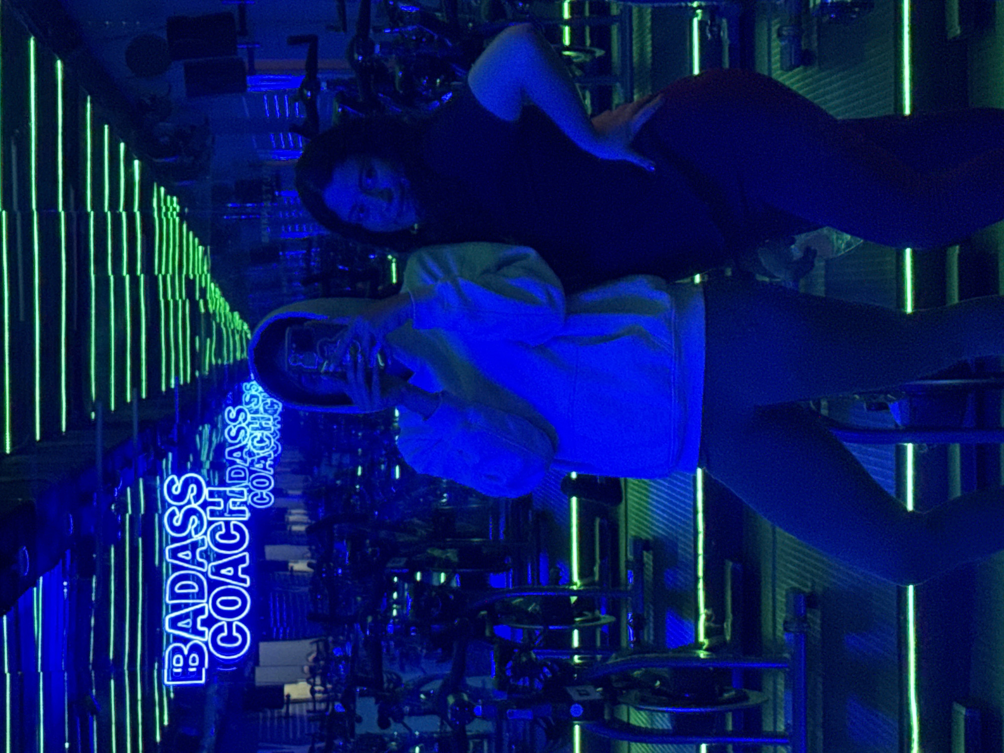 Two individuals are present in a room with neon lighting. One person is wearing a light-colored hoodie and leggings, while the other is in a dark top and leggings. A neon sign reading "BADASS COACH" is visible. The room appears to be a fitness studio, possibly containing exercise equipment. This image was taken inside a gym in Puebla, Mexico.