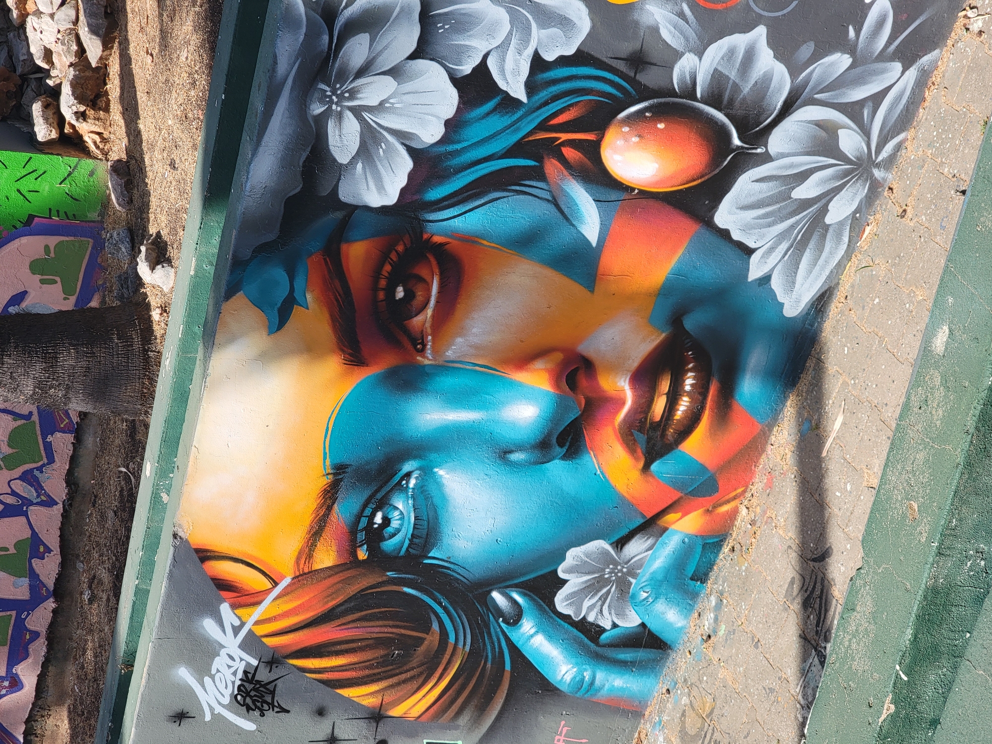 This is a photograph of a vibrant graffiti mural on a concrete wall in Quarteira, Portugal. The mural depicts a stylized portrait of a woman with strikingly contrasting colors. Her face is split down the middle, with one side rendered in shades of blue and orange, and the other in warm oranges and browns. Her eye on the blue side is a piercing blue, while the eye on the brown side is a deep brown. Her lips are full and painted with a dark, glossy lipstick.
The woman's hair is depicted with streaks of orange, yellow, and blue, framing her face. There are also large, white, stylized flowers integrated into the composition, appearing on the right side of her head and near her left hand. A single, glossy orange orb with a drop falling from it hangs near her right cheek. The left side of the mural includes white graffiti lettering, possibly a tag or artist's signature.
The mural is situated outdoors, with a paved walkway in the foreground and a slope leading up to the wall, covered in dry grass and soil. In the background, another section of wall displays more graffiti, this time with larger, abstract shapes in pink, purple, and green. A portion of a tree trunk, possibly a palm tree given the location, is visible behind the mural. The overall impression is one of urban art in a sunny, outdoor environment.