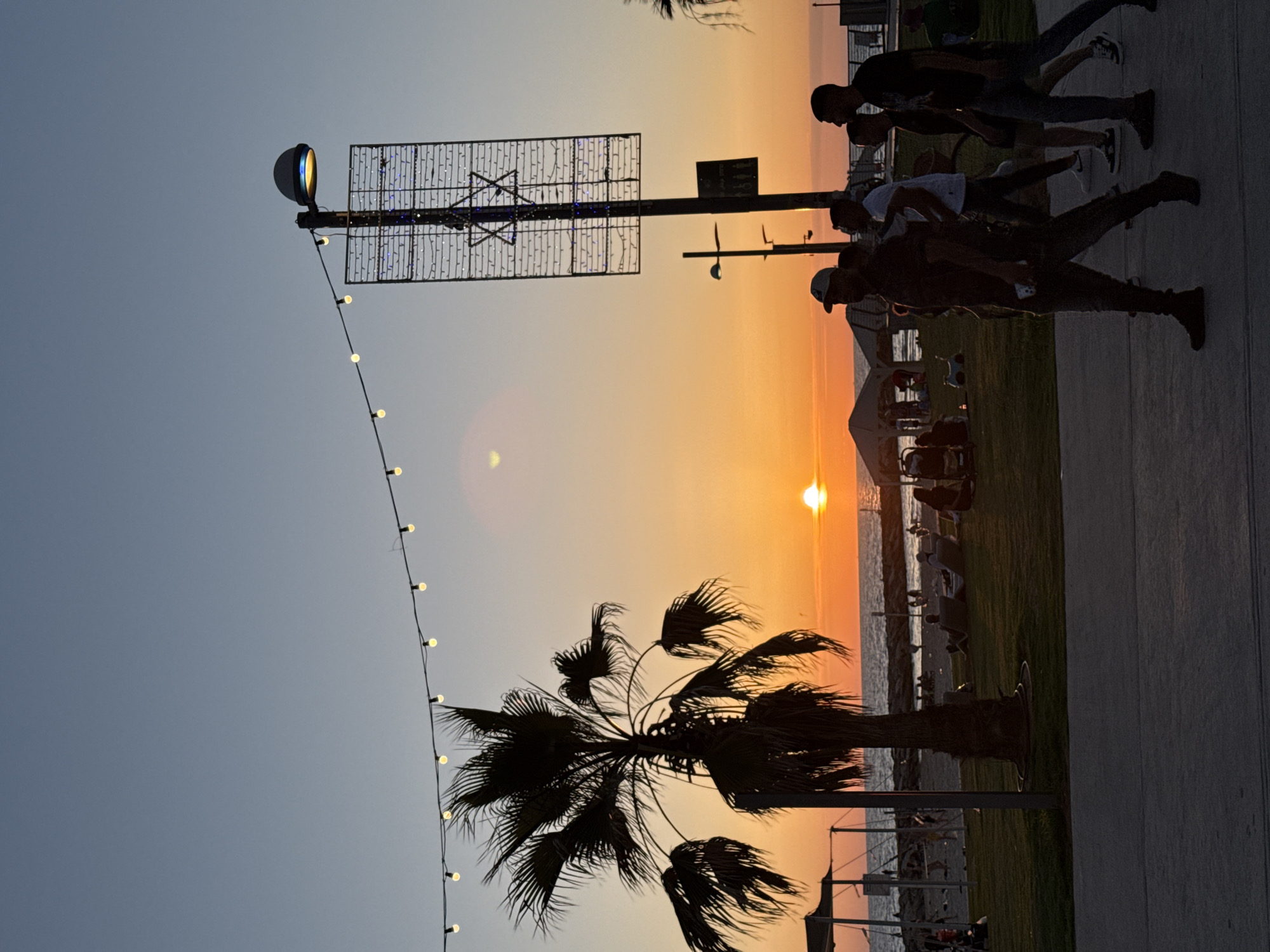 An outdoor scene captured at sunset depicts a vibrant sky over a coastal area in Tel-Aviv, Israel. The sun, a distinct orange disk, is visible on the horizon, casting an intense orange and yellow glow across the lower sky that gradually transitions to a pale blue overhead. A string of illuminated fairy lights stretches horizontally across the upper frame. In the foreground, a dark concrete promenade is visible, upon which several human figures are walking, primarily from left to right. Four male figures are prominently silhouetted on the right; one wears a cap and holds a mobile phone, leading the group. Immediately behind the promenade, a strip of green grass features a large, dark palm tree silhouette. Beyond the grass lies a sandy beach area, occupied by numerous silhouetted individuals relaxing on lounge chairs and on the sand, with several small structures also visible. The Mediterranean Sea stretches across the horizon, containing a rock breakwater extending from the right. On the right side of the frame, a tall pole supports an illuminated street lamp with a rounded white fixture. Attached to this pole is a large rectangular sign, prominently displaying a blue Star of David formed by lights, within a grid of white lights. A circular lens flare is visible in the upper-mid-left sky.