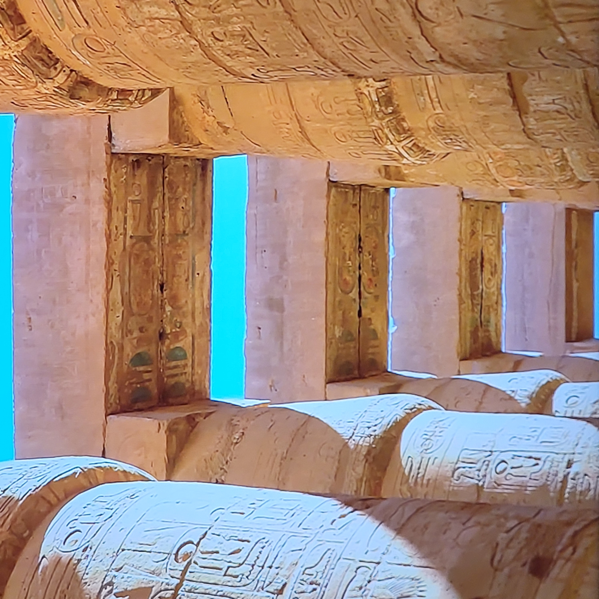 The image shows an interior view of an ancient Egyptian structure. Rows of large, cylindrical columns are visible in the foreground, covered in hieroglyphic carvings. Between the columns, there are glimpses of bright blue sky through rectangular openings. Above the columns, a massive, carved ceiling extends overhead, also decorated with hieroglyphs. There are no people or immediate actions occurring in the scene. The photograph depicts a static architectural space, most likely within a temple or similar structure. The location is not in Yerevan, Armenia, but within Egypt.
