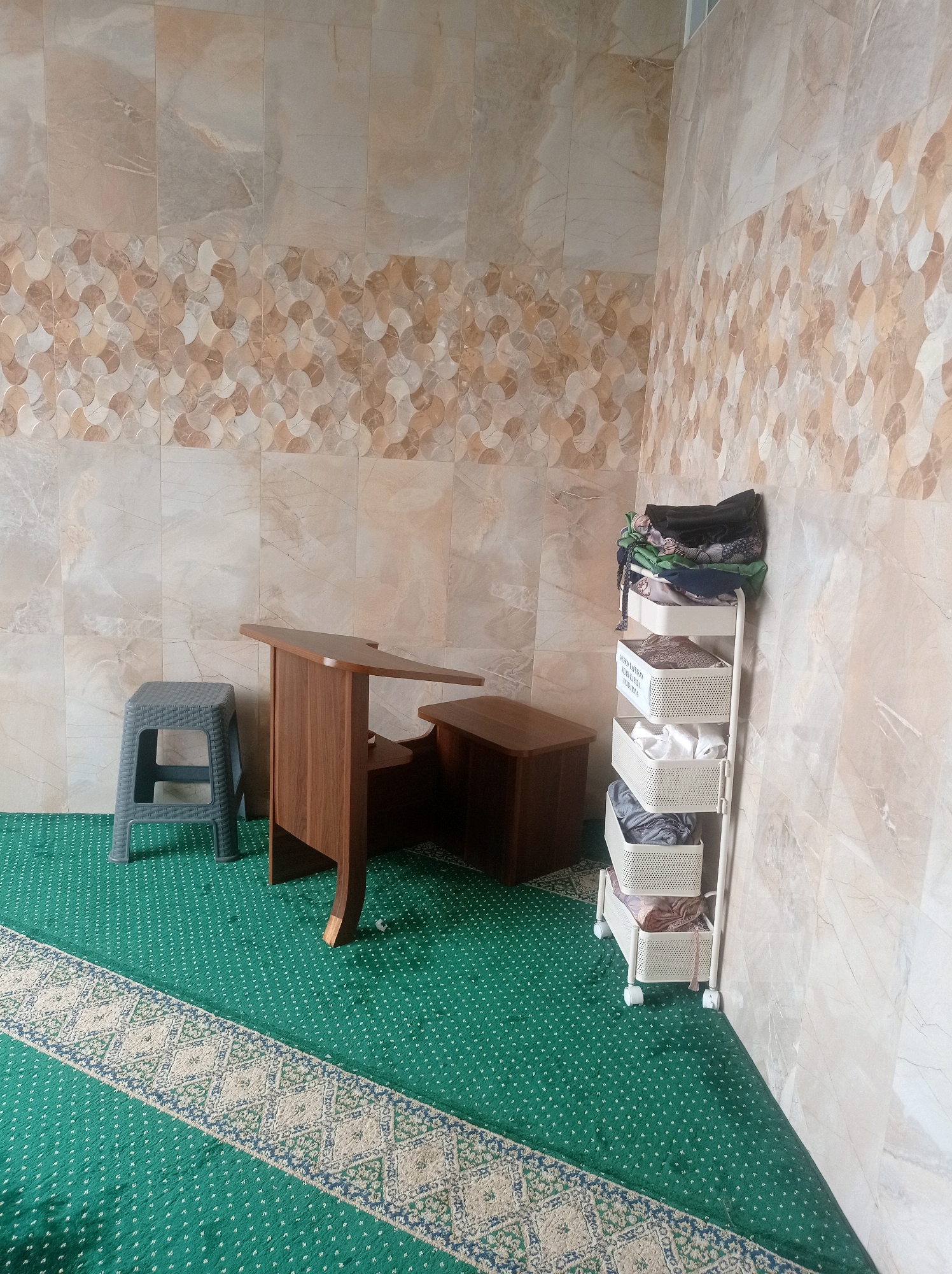 The image depicts a corner of a room, possibly a prayer room or a small study area, with tiled walls and a green carpet. On the left, a grey plastic stool sits next to a wooden desk with a carved edge. To the right of the desk is a smaller wooden cabinet. A white rolling cart filled with folded clothes and textiles stands against the wall on the right. The wall tiles are a neutral tone with a decorative border of circular patterns. The green carpet has a repeating small white pattern and a decorative border with diamond shapes. The overall impression is of a functional, clean, and modestly furnished space. There are no people visible in the image, and no specific location in Bandung City, Indonesia can be identified from the visual cues alone.