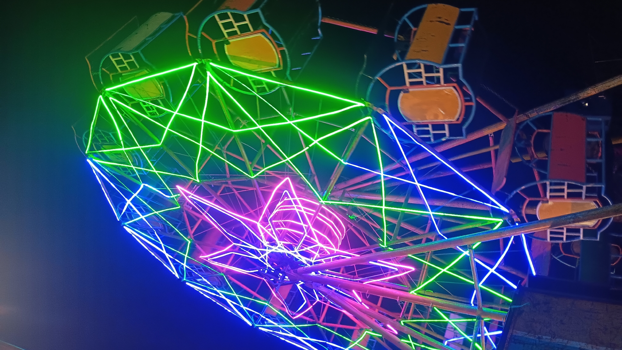 A Ferris wheel is illuminated by neon lights in the city of Getas, Indonesia. The Ferris wheel is decorated with blue, pink, and green neon lights. The lights form geometric patterns across the structure of the wheel. The sky is dark, indicating that it is nighttime. The bottom of the Ferris wheel is visible, with some of its cabins also illuminated. The image is taken from a low angle, looking up at the Ferris wheel.