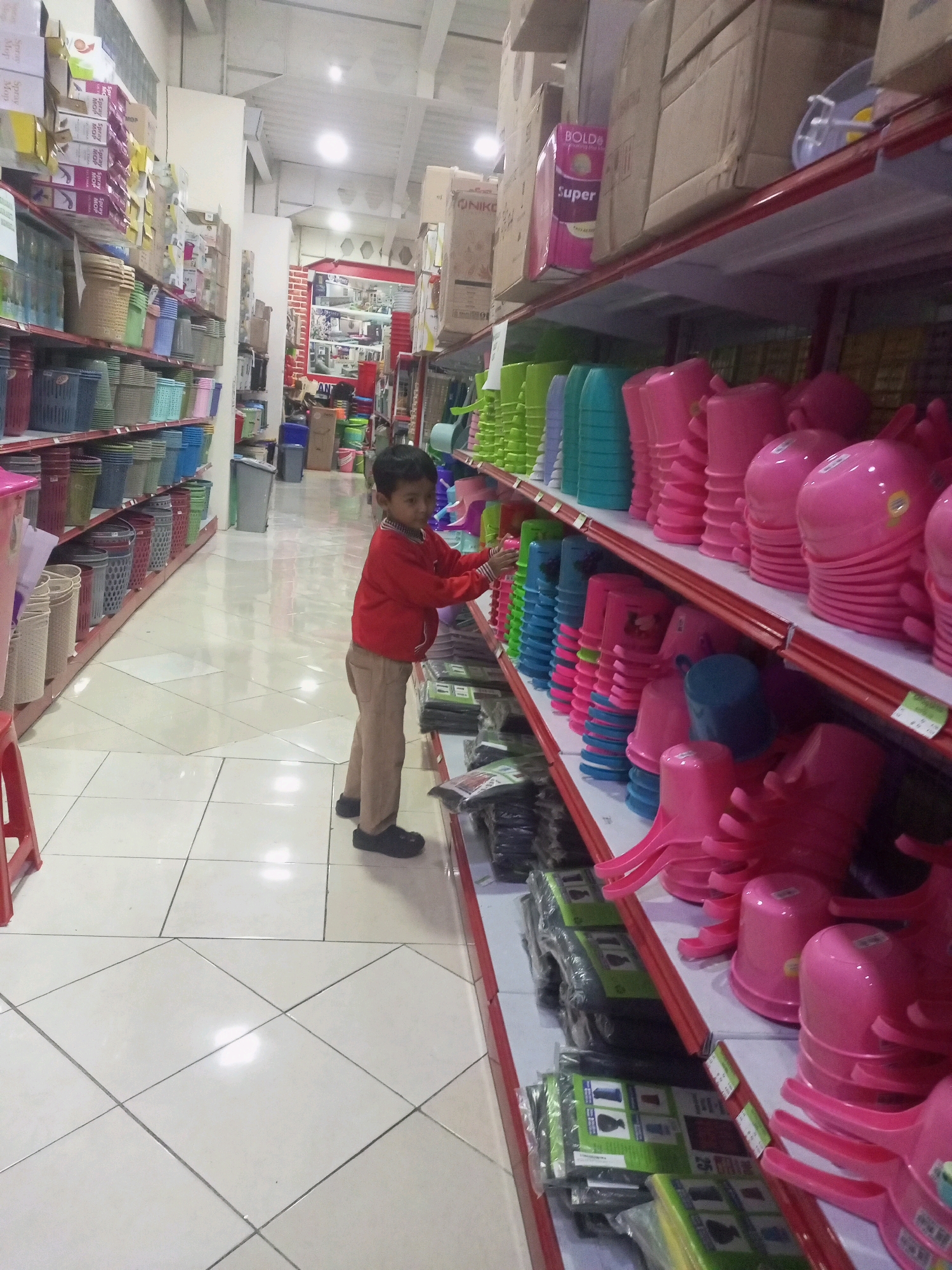 In a retail store aisle in Ngasem, Indonesia, a young boy, wearing a red jacket and tan pants, stands between two shelves stacked with household plastic goods. The shelves on the left are filled with various sizes and colors of baskets and containers, while the shelves on the right are predominantly stocked with pink and green plastic items, including what appear to be buckets or similar containers with handles. The store's floor is tiled with large, light-colored square tiles with dark grout lines, reflecting the overhead fluorescent lighting. Cardboard boxes are visible on higher shelves and in the background, suggesting a large inventory. The overall environment appears to be a well-lit, organized shop selling plastic housewares.