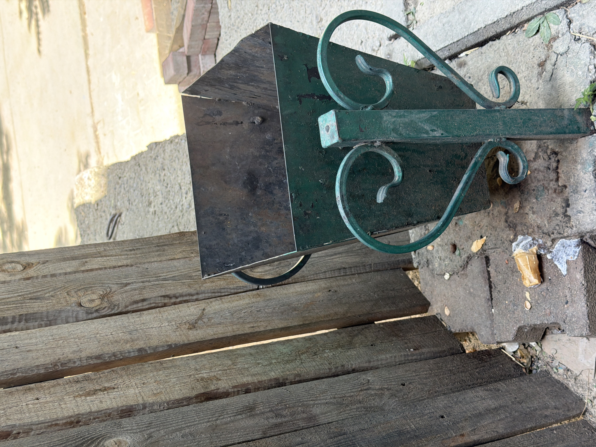 This is a medium-shot, eye-level, outdoor photograph of a green metal trash can situated next to a weathered wooden fence. The trash can has a decorative green metal scrollwork base with two heart-shaped elements. The can itself appears to be made of dark metal, with visible wear and some residue inside. To the left of the trash can, vertical wooden planks form a fence, showing signs of age and exposure to the elements. On the ground in front of the trash can, there is some debris, including what appears to be a discarded ice cream cone. The ground surface is a mix of concrete and gravel. In the background, a paved area and a pile of bricks are visible. The overall scene suggests an urban or semi-urban setting in Shymkent, Kazakhstan, likely a public space such as a park or street. There are no people visible in the image.