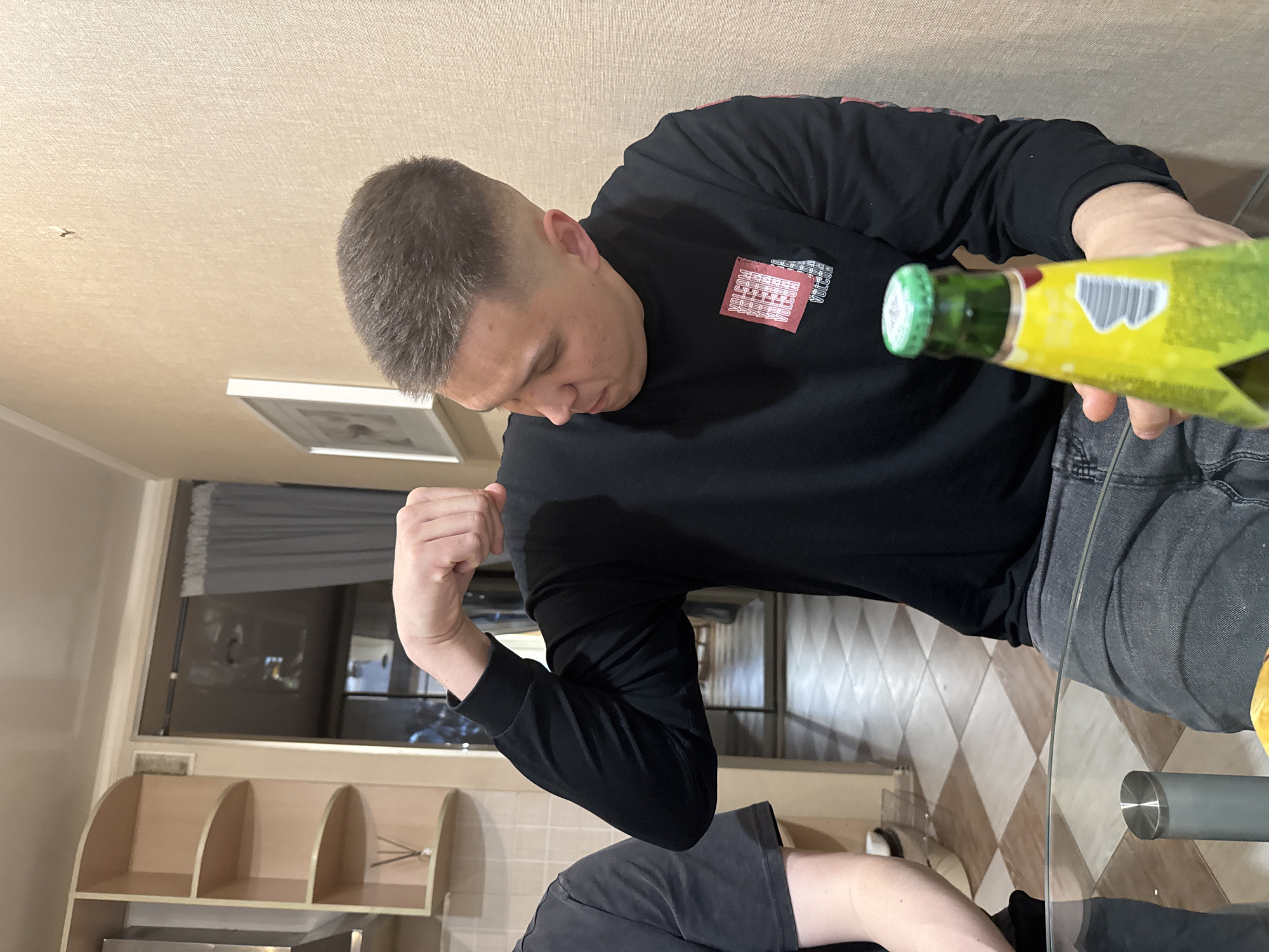 A man in a black long-sleeved shirt and grey jeans is seated at a glass table in what appears to be a kitchen or dining area. He is holding a yellow and green bottle, and his other arm is flexed, showing his bicep. The background includes a wall with light brown wallpaper, a window with curtains, and kitchen cabinets. Based on the provided information, the location is Kazan, Russia. The flooring is a diamond pattern of light and dark brown tiles. Another person wearing a black shirt is visible partially at the bottom of the frame.