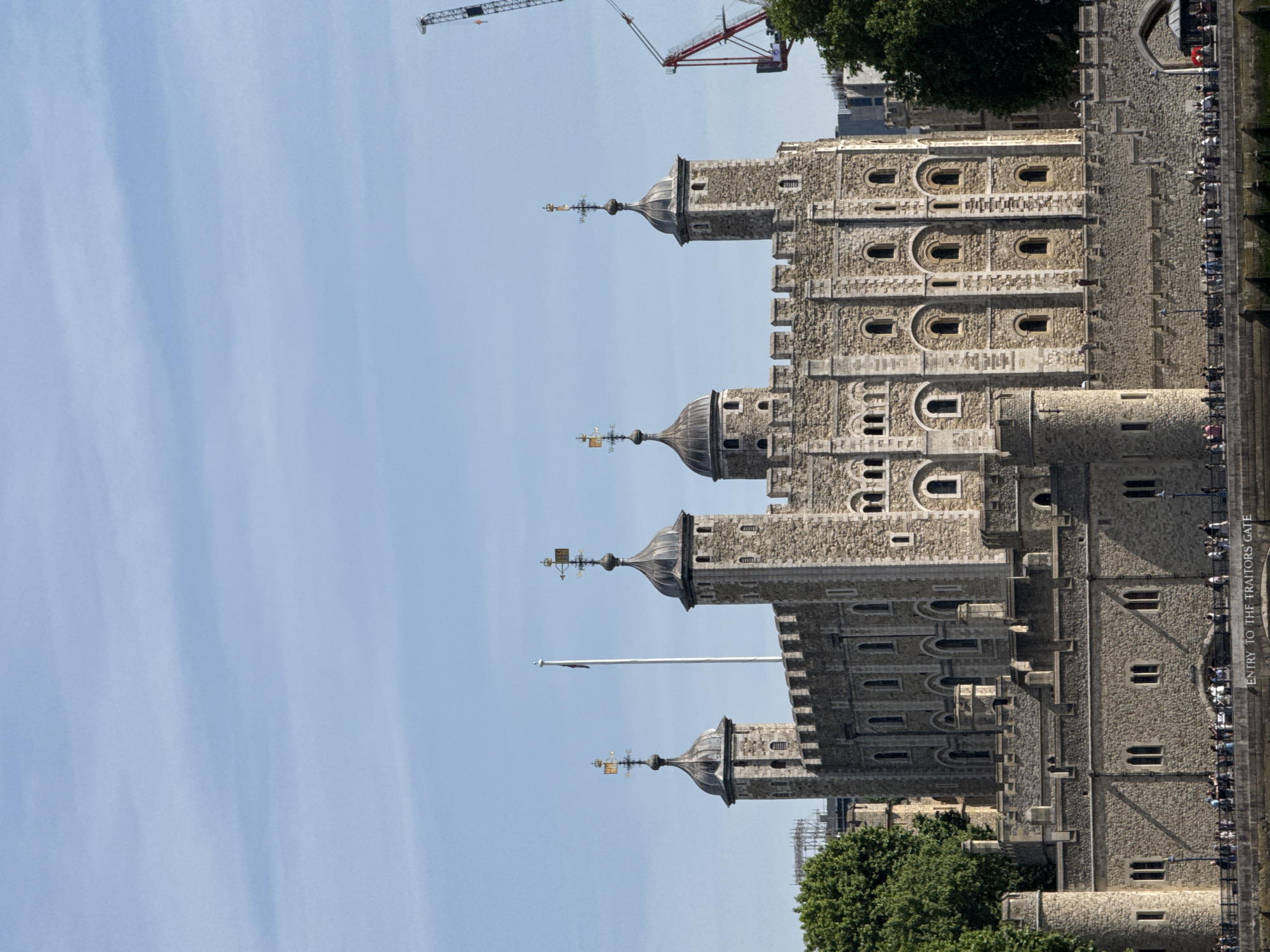 The image captures a view of the Tower of London in the city of London, United Kingdom. The architecture of the historic castle complex is prominently featured, displaying its aged stone walls and distinctive towers with turrets. In the background, glimpses of modern construction cranes indicate ongoing development in the area. On the side of the tower, there is a crowd of people walking along the path that is labelled "Entry to the Traitors Gate." The sky is clear and blue.