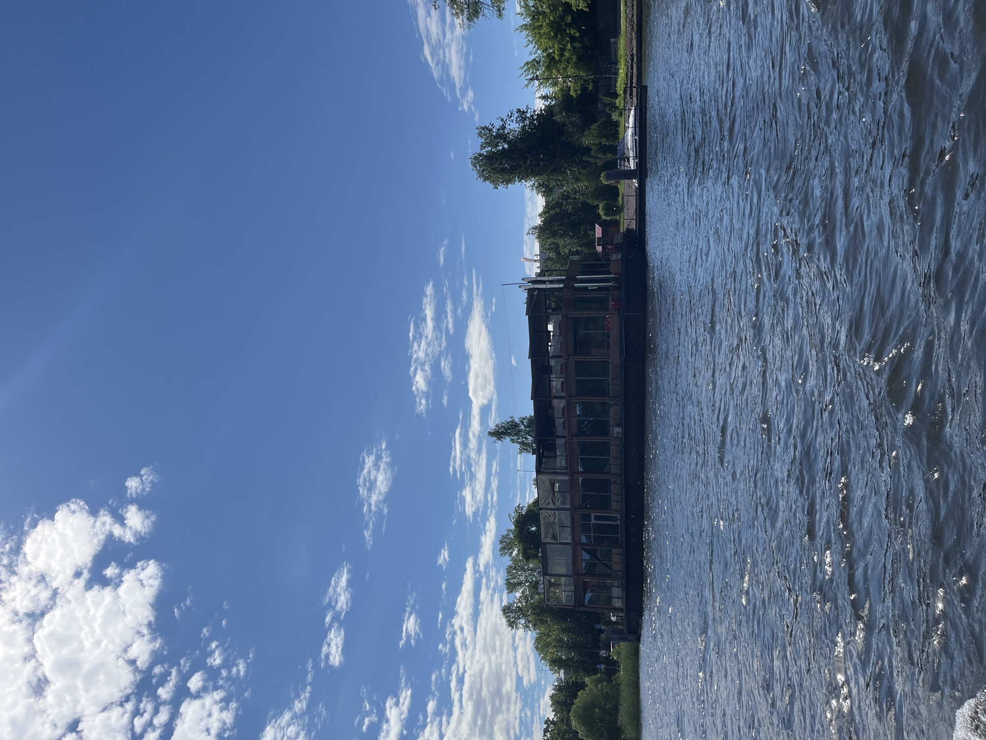 A vertical, wide shot captures a serene scene on a body of water, likely a river or canal, in Saint Petersburg, Russia. The water, reflecting the bright blue sky and scattered white clouds, ripples gently, creating a shimmering effect. Along the right bank, a modern, multi-story structure with a dark facade and numerous large windows is docked. The building appears to be some sort of establishment, possibly a restaurant or event venue, with an industrial aesthetic.
Lush green trees line the bank behind the structure, suggesting a park-like or natural setting. A few bare vertical poles, possibly part of the building's infrastructure or related to nearby navigation, are visible. The clear blue sky indicates it's daytime, likely late morning or afternoon, with pleasant weather. The overall mood is peaceful and tranquil, evoking a sense of calm and leisure. There is no visible text in the image.