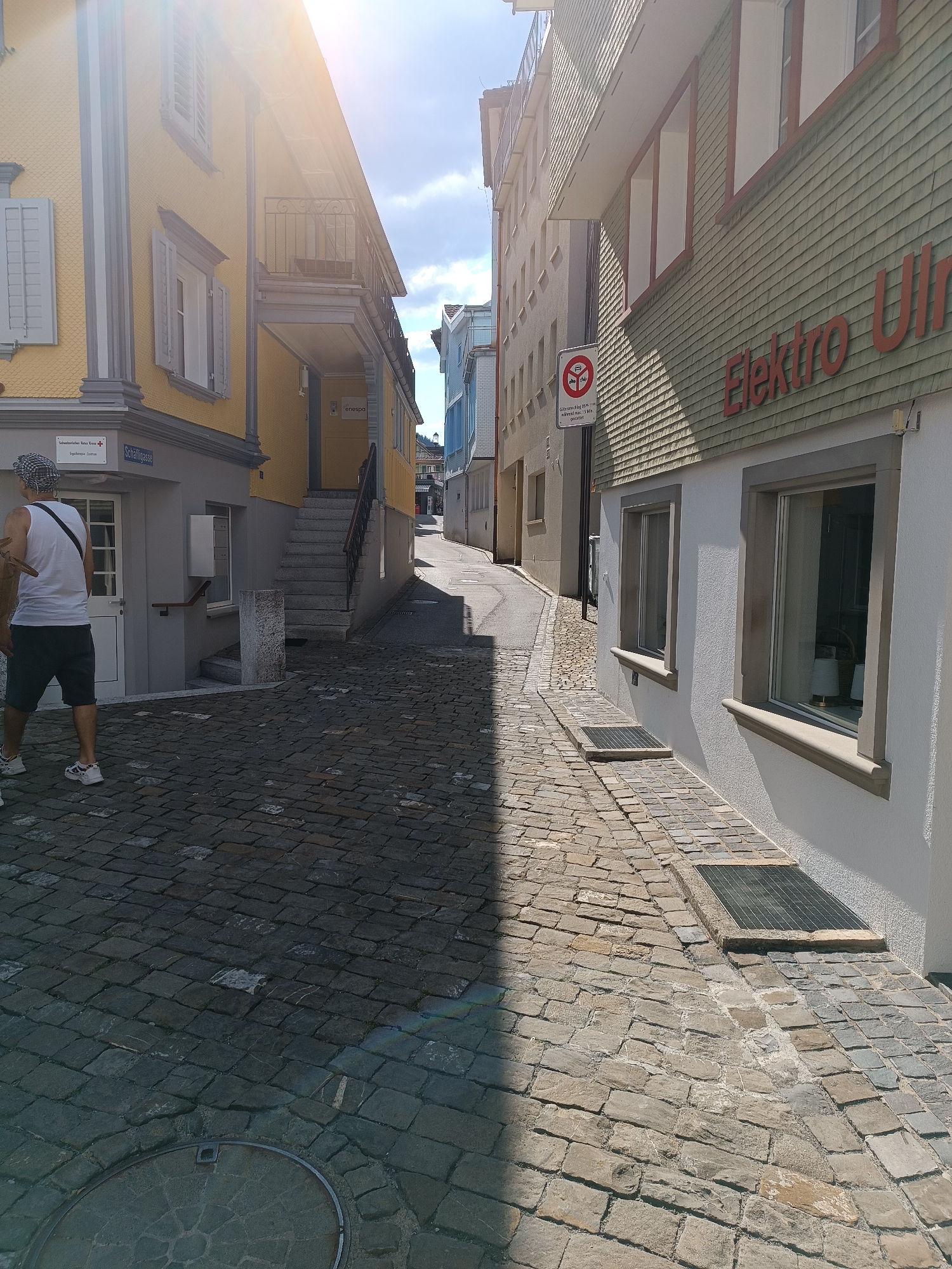 The image shows a narrow, cobblestone street in Appenzell, Switzerland, lined with traditional buildings. On the left, a yellow building with grey trim features windows with shutters and a balcony. A sign on this building reads "Schweizerisches Rotes Kreuz" (Swiss Red Cross). Further down the street, a person wearing a white tank top, dark shorts, and a checkered hat is walking away from the camera.
On the right side of the street, a building with a light green, shingled facade displays large red lettering that appears to say "Elektro Uhren" (Electric Clocks). Below this, a window is visible. Further down the street, a red and white traffic sign is posted, indicating restrictions for motorized vehicles.
The street is paved with uneven cobblestones, and shadows from the buildings create a stark contrast across the scene. The sky is bright with sunshine and some clouds. The overall impression is of a historic European town street.