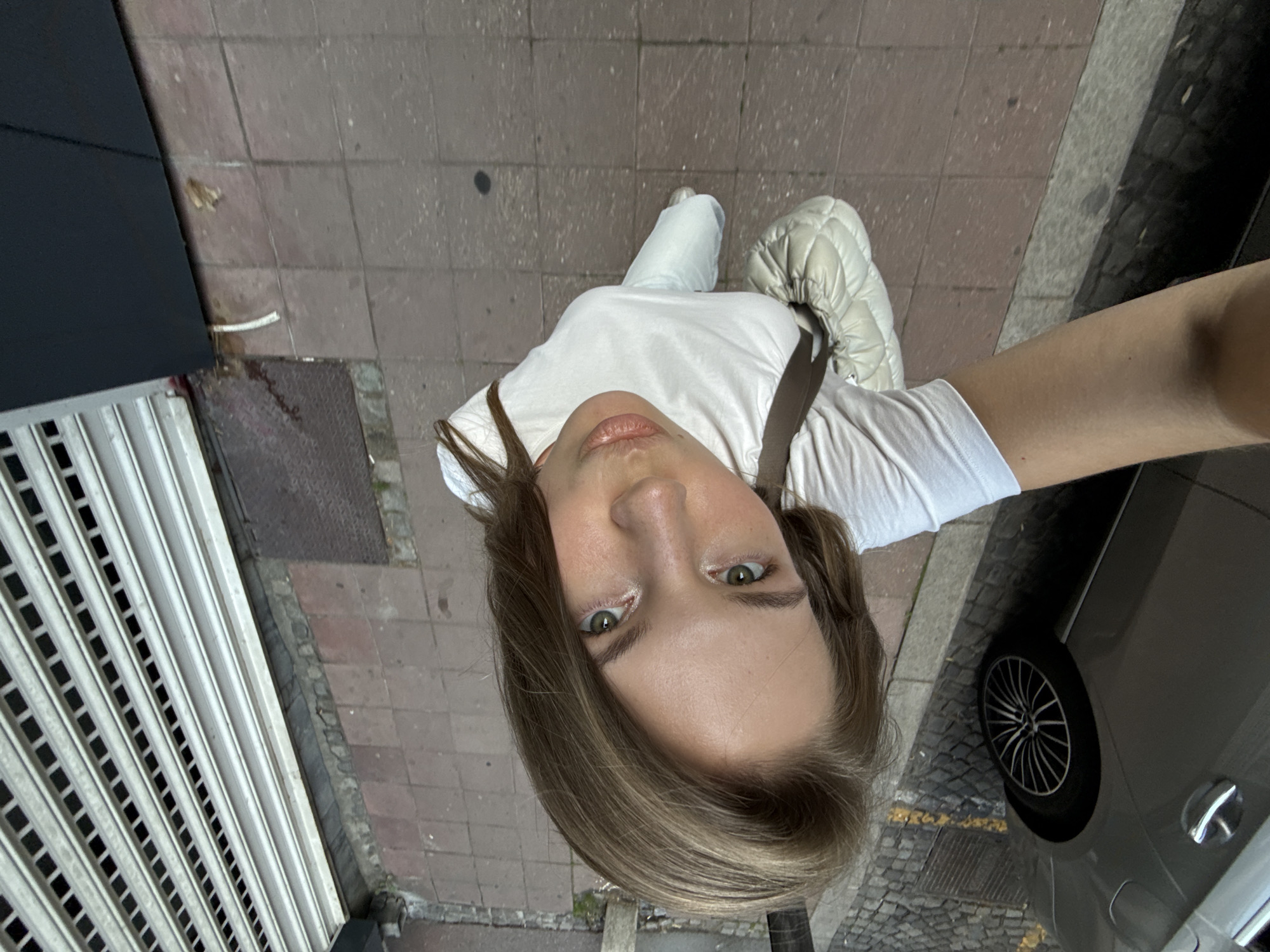 A first-person perspective selfie shows a young woman with light brown hair and green eyes, wearing a white t-shirt and white pants. She is standing on a city sidewalk with red-brown square tiles. To her left, a metal roll-up gate is partially visible. In the foreground, the top of a gray car and its wheel are seen. Behind her, the sidewalk continues, with cobblestones on the far left and a metal grate. The image is taken from a high angle, looking down at the woman. The scene appears to be outdoors in an urban environment. The location is identified as Belgrade, Serbia.