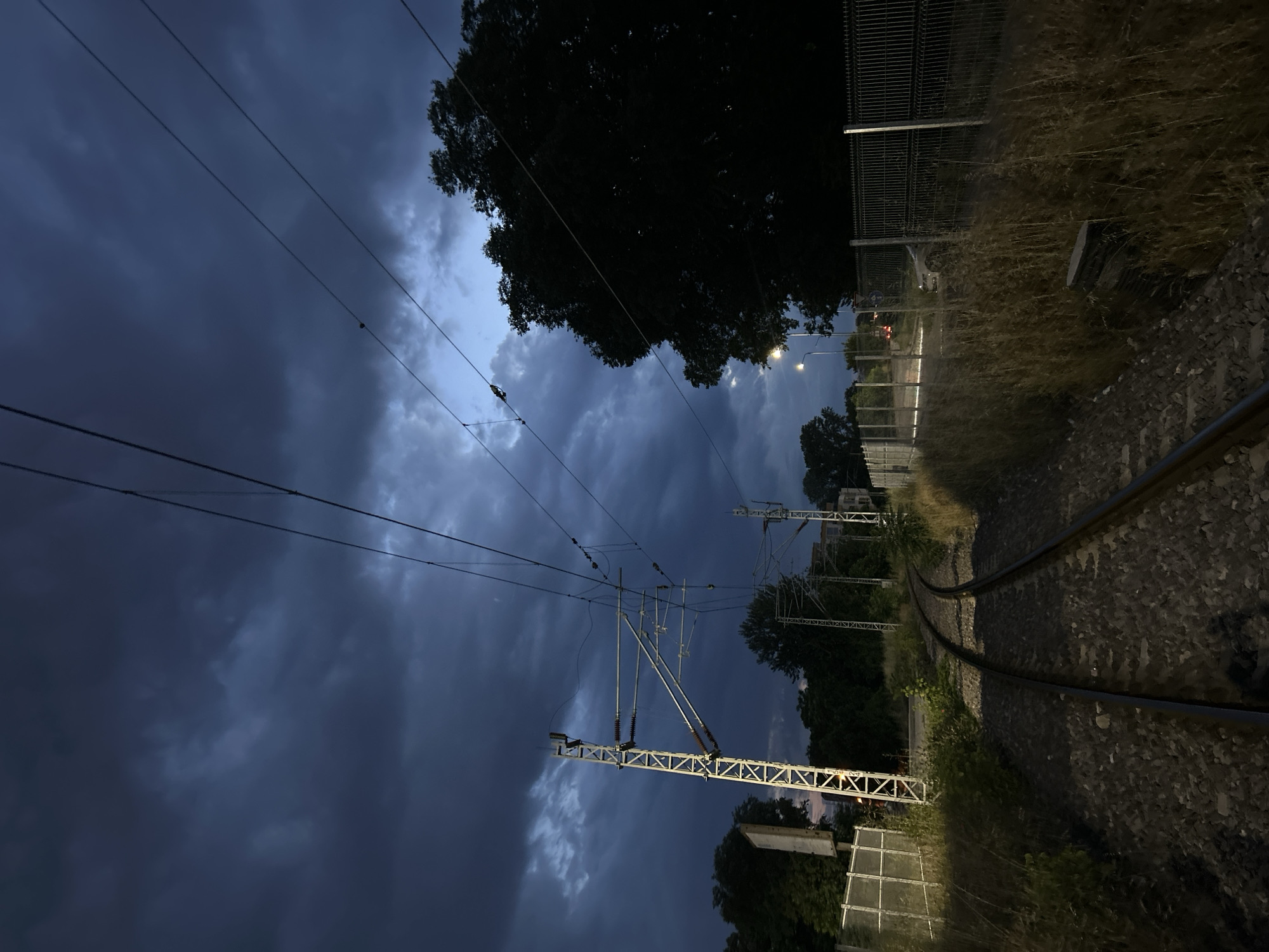 The image depicts a railway scene in Plovdiv, Bulgaria, captured during late evening or twilight under a heavily clouded sky. The dominant feature is the dark, dense cloud cover that fills the upper portion of the frame, showing only faint patches of lighter sky. Below this, a complex overhead catenary system for railway electrification spans the tracks, with numerous wires and robust metal support structures, including a prominent mast on the left side, equipped with insulators.
In the foreground, a set of railway tracks curves gently towards the right and recedes into the distance. The ground alongside the tracks on the right is covered with tall, dry grass and weeds, while a metal mesh fence runs parallel to the track. Further along the right side, multiple artificial light sources, likely streetlights, cast a soft, warm glow, illuminating sections of the fence and the ground. Dense, dark green trees with thick foliage are silhouetted against the sky, forming a prominent line of trees on the right and in the background.
The overall atmosphere is quiet and somewhat moody due to the dim lighting and heavy clouds. No people or specific activities are visible, emphasizing the industrial infrastructure and natural elements. No discernible text is visible within the image.