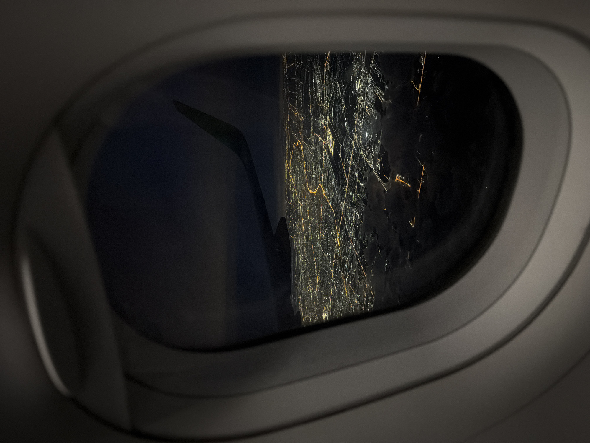 A view from an airplane window shows a cityscape at night. The illuminated city lights resemble a grid-like network of streets and buildings. The wing of the plane is visible in the left portion of the window. Based on the style of urban lighting, the location is likely Bangkok, Thailand.