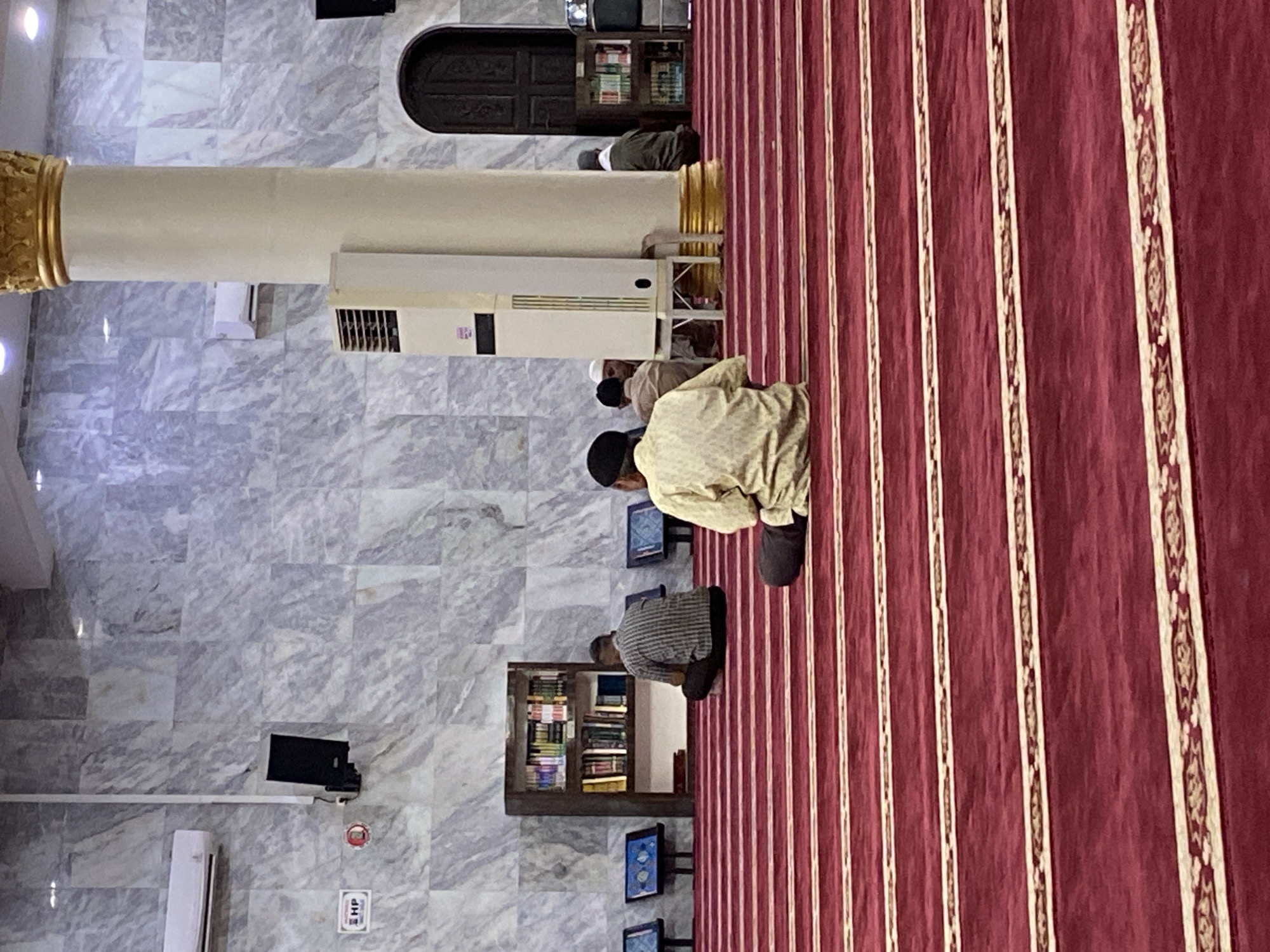 This image depicts the interior of a mosque, specifically the prayer hall, in Lhokseumawe, Indonesia. The foreground is dominated by a rich, red carpet with gold and white striped patterns, indicating designated prayer rows. Several individuals are seated on the carpet, facing away from the camera, engaged in prayer or contemplation.
In the middle ground, a man in a light-colored, patterned garment is seated in the center, facing away from the viewer. To his left, another man in a striped shirt is seated. Further back, near a bookshelf filled with religious texts, another individual is visible. A tall, white pillar with a golden capital stands prominently to the right of the frame, supporting the mosque's ceiling.
The walls are adorned with marble tiles, and in the background, a dark wooden door is visible. Air conditioning units are mounted on the walls, and a speaker is attached to the wall near an "HP" sign. The overall environment suggests a place of worship, likely during a prayer service or a period of personal devotion. The lighting appears to be artificial, casting a warm glow on the scene.