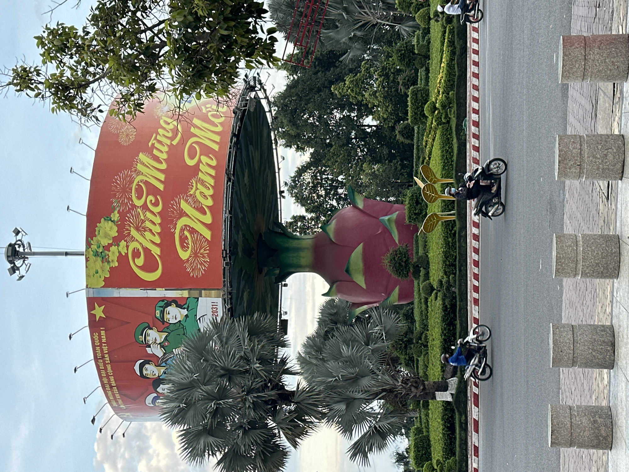 A cylindrical billboard displaying a Vietnamese New Year greeting "Chúc Mừng Năm Mới" is supported by a large, stylized dragon fruit structure. The billboard features a red background with yellow text and images of people in green uniforms and a yellow star, along with fireworks. To the left of the billboard, a tall palm tree with fan-shaped fronds stands out. In the foreground, a two-lane road runs horizontally across the image. Two individuals on motorcycles are riding on the road. The motorcycle on the left is white and ridden by a person wearing a blue shirt and a helmet. The motorcycle on the right is black and ridden by a person wearing a dark jacket and a face mask. A median strip with red and white stripes separates the lanes of traffic. Along the roadside, there are meticulously manicured green hedges and shrubs. In the immediate foreground, three cylindrical concrete bollards are positioned on a paved walkway. The scene appears to be an outdoor urban setting with a focus on a prominent public display and traffic. The billboard text "Chúc Mừng Năm Mới" translates to "Happy New Year" in Vietnamese, and the surrounding imagery suggests a festive, celebratory occasion, likely associated with the Lunar New Year. The presence of uniformed figures and communist symbols on the billboard indicates a patriotic and political theme. The dragon fruit sculpture is a unique architectural element that integrates a local produce with the advertising structure.
