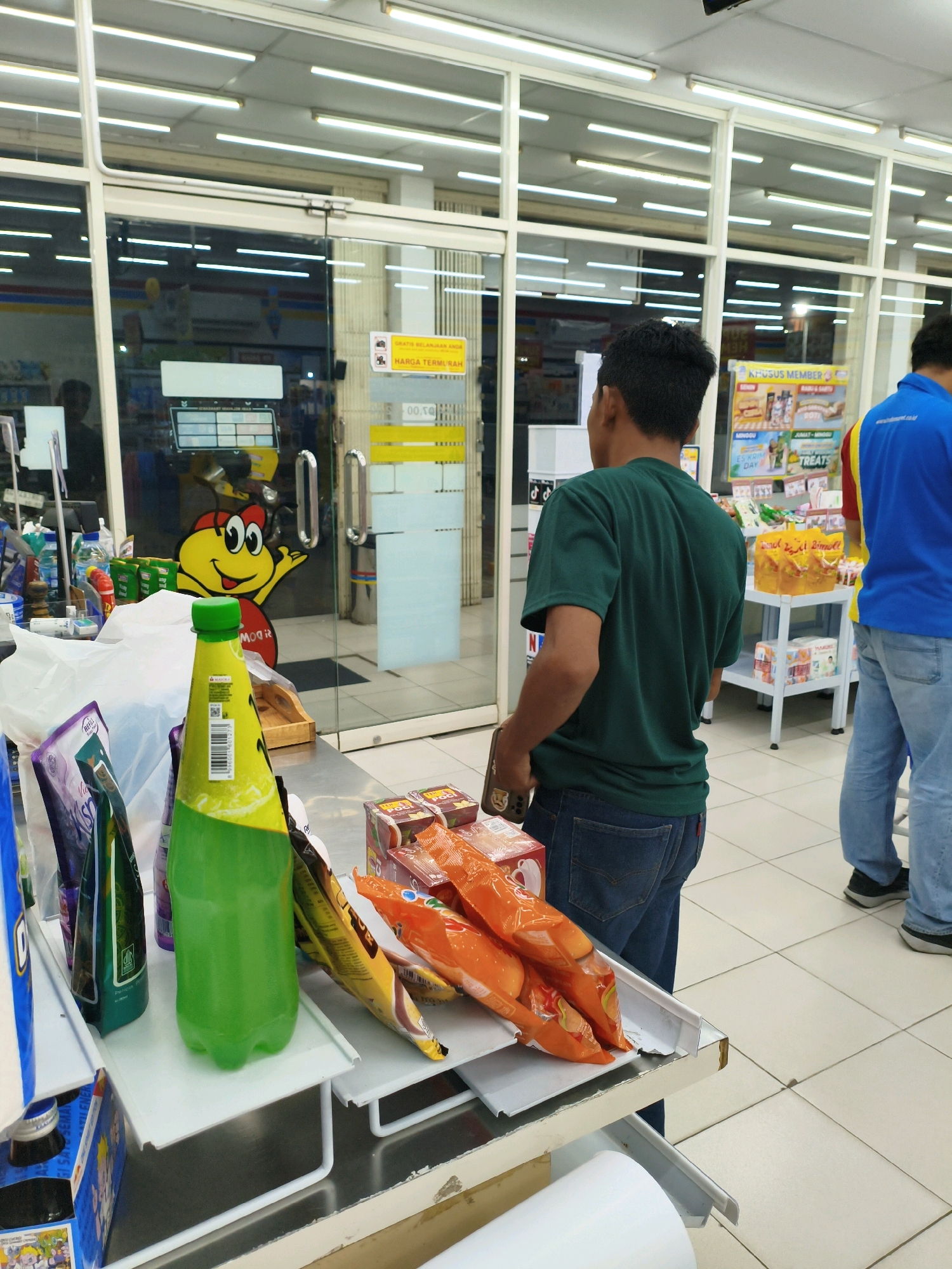 The image depicts the interior of a convenience store, likely a branch of Indomaret or Alfamart given the typical store layout and product placement seen in Indonesia. The scene is set at a checkout counter, with a young man in a green t-shirt and blue jeans standing with his back to the camera, appearing to be paying for items. In the foreground, on the counter, are various products including a large green bottle of drink, several bags of snacks (some orange and some with purple packaging), and a box of what appear to be chocolates or candies. Behind the counter, a cashier's station is partially visible. The store's interior is brightly lit with fluorescent ceiling lights. Through a glass door, other shoppers can be seen, and a cartoon character decal is visible on the door. The floor is tiled in a light color. While the specific location is given as Sumberejo Wetan, Indonesia, the image itself doesn't contain any explicit signage or landmarks that would confirm this location beyond what's provided. The overall impression is a typical transaction in a common Indonesian retail environment.