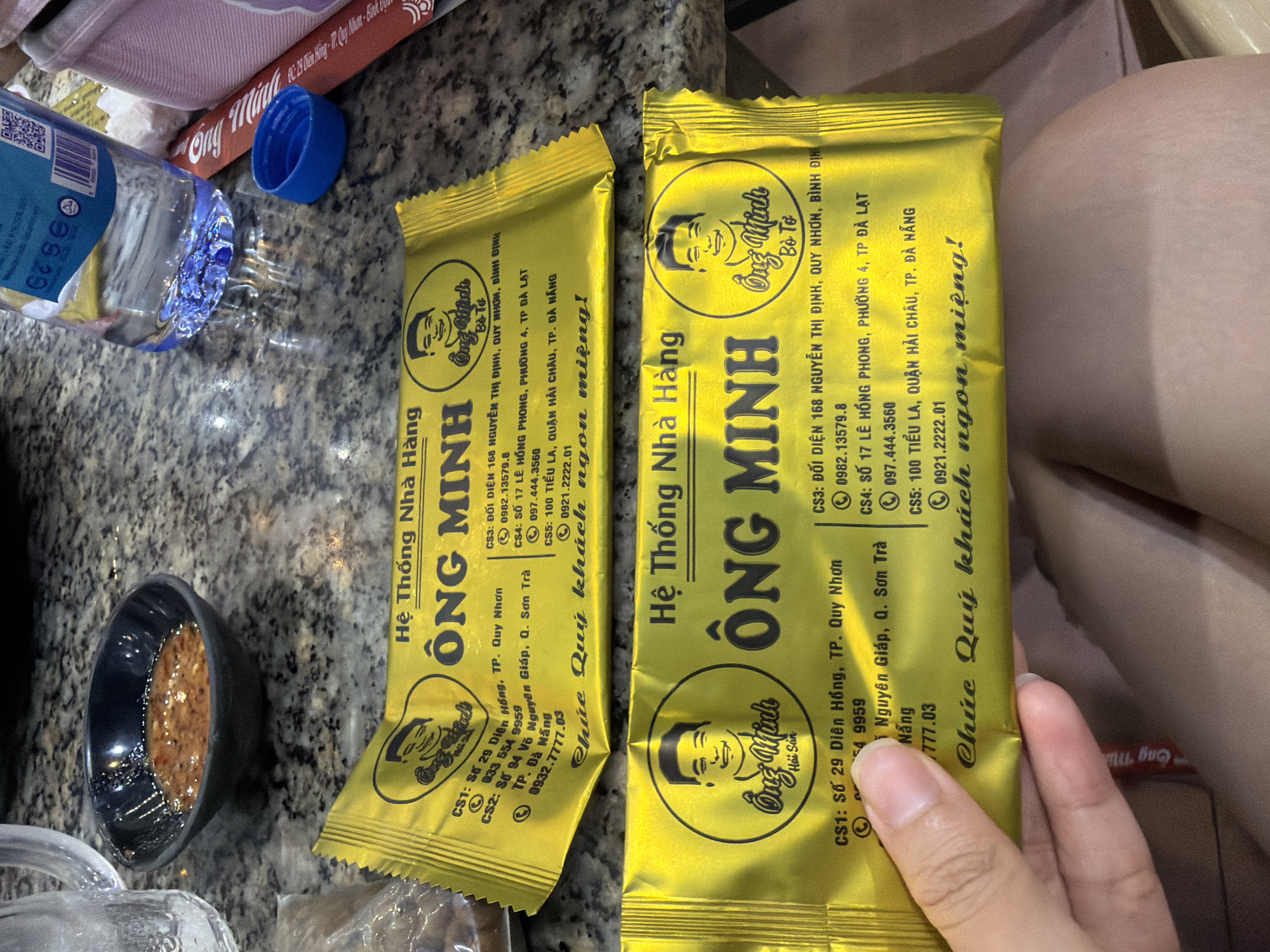 The image displays a tabletop scene featuring two golden-yellow packaged food items, a small bowl of sauce, and a water bottle, suggesting a snack or meal setting. A person's hand is visible on the right, holding one of the packages.
The primary subjects are two identically branded, crinkled golden-yellow pouches from "Hệ Thống Nhà Hàng ÔNG MINH" (Ông Minh Restaurant System). Both feature a cartoon illustration of a man's face. Below the man's face, one package reads "Bò Tơ" (Young Beef) and the other "Hải Sản" (Seafood). A slogan, "Chúc Quý khách ngon miệng!" (Wishing you a delicious meal!), is prominently displayed. To the left of the packages is a small black bowl containing a brown, textured dipping sauce. A clear plastic water bottle, lying on its side with a partially visible label including a QR code, and a detached blue plastic bottle cap are also on the table. The items rest on a grey, speckled countertop. In the blurred background, a pink or light red object is visible.
The packages list multiple branch addresses and phone numbers in Vietnam, including:
- CS1: 29 Dien Hong, Quy Nhon City
- CS2: 94 Vo Nguyen Giap, Son Tra District, Da Nang (phone: 0932.7777.03)
- CS3: Opposite 168 Nguyen Thi Dinh, Quy Nhon, Binh Dinh (phone: 0982.13579.8)
- CS4: 17 Le Hong Phong, Ward 4, Da Lat City (phone: 097.444.3560)
- CS5: 100 Tieu La, Hai Chau District, Da Nang City (phone: 0921.2222.01)
These location cues align with the context of Quy Nhon Ward, Vietnam. The time of day is not discernible due to indoor lighting. No specific emotions are conveyed.