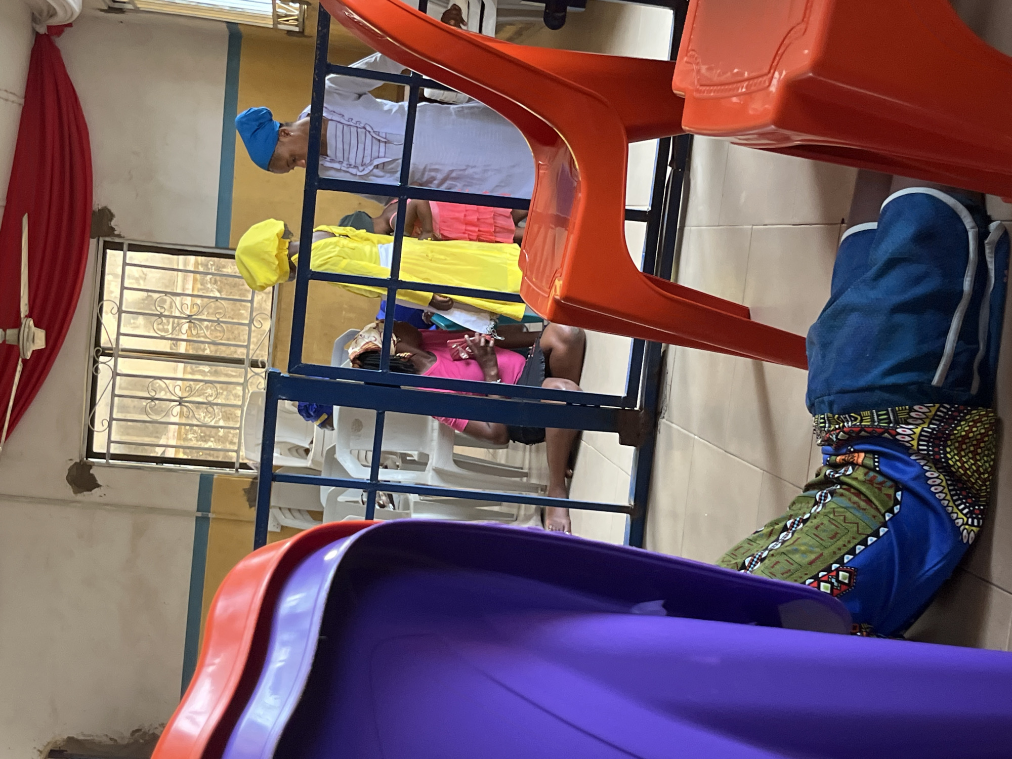 The original image is captured from a low angle, looking upwards and across an indoor space, and is rotated 90 degrees counter-clockwise.
In the immediate foreground, a stack of brightly colored plastic chairs, primarily purple and red, is partially visible at the bottom right (when viewed upright). Just behind these chairs, a person's legs are visible, wearing blue trousers with a vibrant, multi-colored patterned fabric (likely a traditional wrap or skirt) in shades of green, blue, yellow, and red, draped over them.
A blue metal barrier or railing structure extends horizontally across the midground, separating the foreground from a group of seated individuals. Through this structure, several people are visible. One person, positioned closer to the foreground, is wearing a bright pink top and dark shorts, with bare feet, and appears to be looking down at something held in their hands. Above them, another individual wears a yellow tunic or dress paired with a matching yellow head covering. Further back and slightly higher, a third person is dressed in a white garment with vertical dark stripes and a blue head covering. Partial views of other individuals, including one with a green head covering, are also discernible. All visible individuals are seated on white plastic chairs behind the metal barrier.
The background shows interior architectural details: a light-colored wall featuring a horizontal blue stripe. A window fitted with an ornate metal security grille is visible on the left, adjacent to a hanging red curtain. The floor throughout the space is covered with light-colored tiles. The scene appears to depict an indoor gathering or waiting area, possibly in a community center, clinic, or similar public space. No discernible text is visible in the image.
