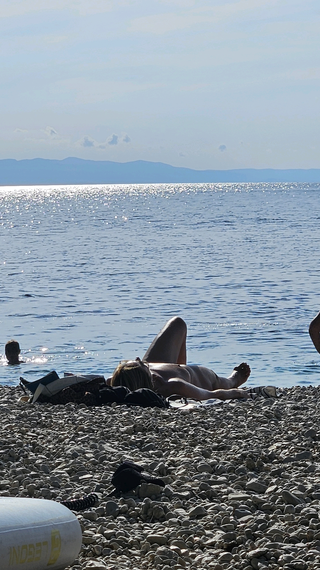 The image depicts a beach scene in Općina Podgora, Croatia. In the foreground, a person is lying on a pebble beach, facing away from the camera, with their body partially visible. They are tanned and appear to be sunbathing. Several items of clothing and bags are scattered on the beach near the person. In the background, the blue sea stretches out, with gentle waves and sunlight glinting on the surface. In the mid-ground, another person's head is visible above the water, suggesting they are swimming or wading. Further in the distance, a hazy mountain range is silhouetted against the sky. The overall impression is a relaxed and sunny day at the coast.