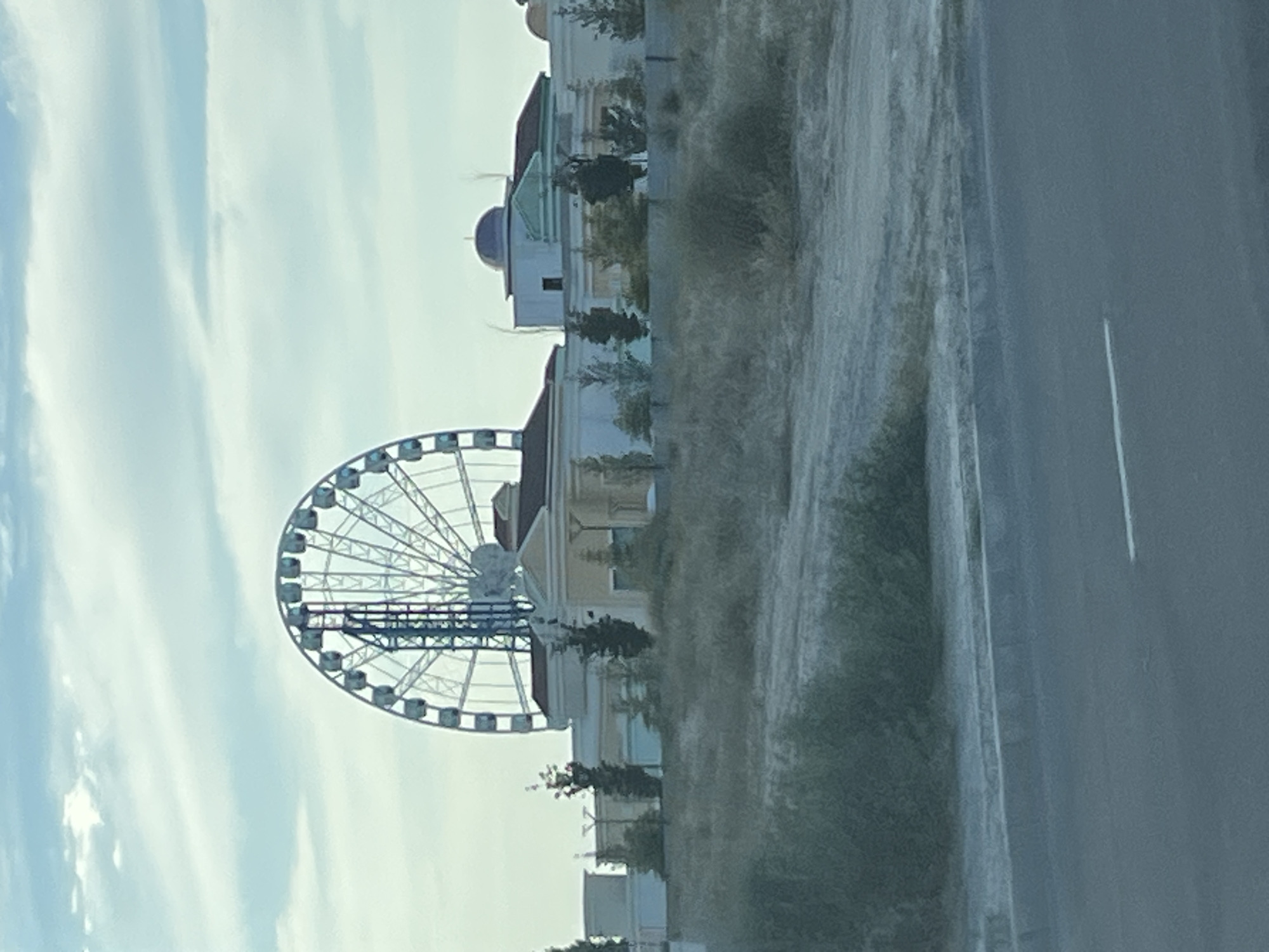 The image shows a Ferris wheel and a modern-looking building complex with a domed structure. The Ferris wheel is large and white with numerous gondolas. The building complex appears to be a commercial or entertainment venue, with a classical architectural style, featuring columns and a reddish-brown roof. The area in front of the building is undeveloped and covered with dry vegetation and dirt. A road is visible in the foreground, with a white line painted on it. The sky is partly cloudy, with a pale blue hue. This scene is located in the city of Aktau, Kazakhstan, likely in a newly developed or developing area, given the contrast between the modern structures and the uncultivated land. There are no people visible in the image.