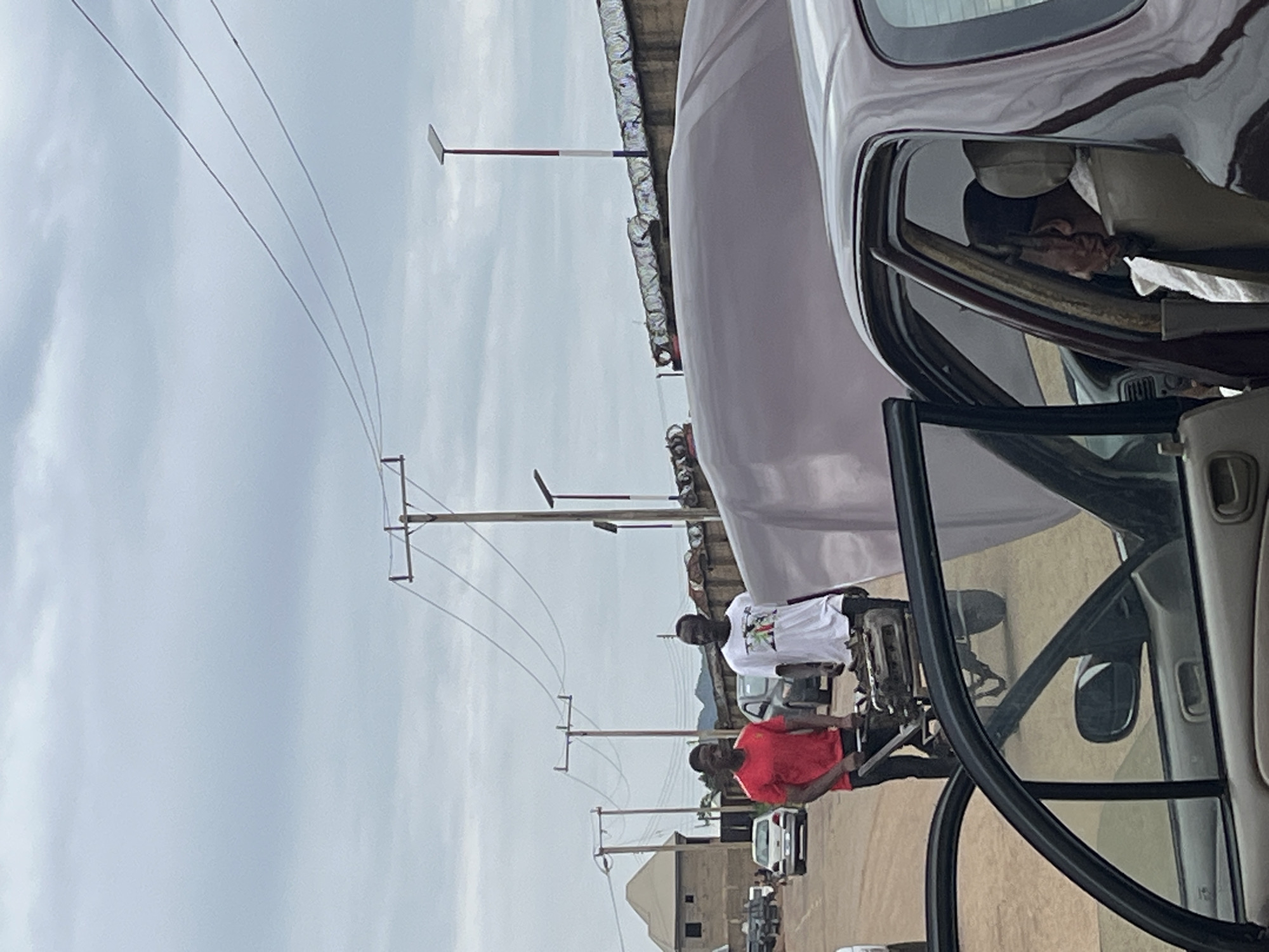 The image shows an outdoor scene in Rafin Par, Nigeria. Two men are walking in the foreground, one wearing a red shirt and the other a white t-shirt, pushing a wheelbarrow that appears to contain mechanical parts. In the middle ground, several cars are parked and driving on a dirt road. A large, indistinct object, possibly a vehicle or structure covered with a tarp, is present. In the background, there are buildings, telephone poles with wires, and a fence topped with barbed wire. The sky is overcast. The perspective is from inside another vehicle, with its door open and partially visible in the foreground.