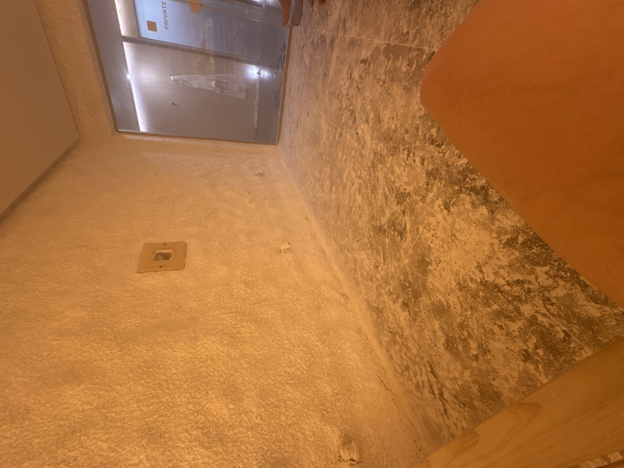 The image shows a section of a room, possibly a bathroom or spa-like area, with a focus on the wall and floor. The wall has a textured, off-white surface with a rectangular electrical outlet. The floor is tiled with a pattern of light and dark shades. A portion of a person's leg is visible in the foreground. A window or glass partition with "PRIVATE" printed on it is at the top left, and through the window, a glimpse of an interior space is visible. The presence of a wooden beam along the bottom suggests the location may be a sauna or similar space. The location in the city of Kazanlak, Bulgaria cannot be determined from this image.