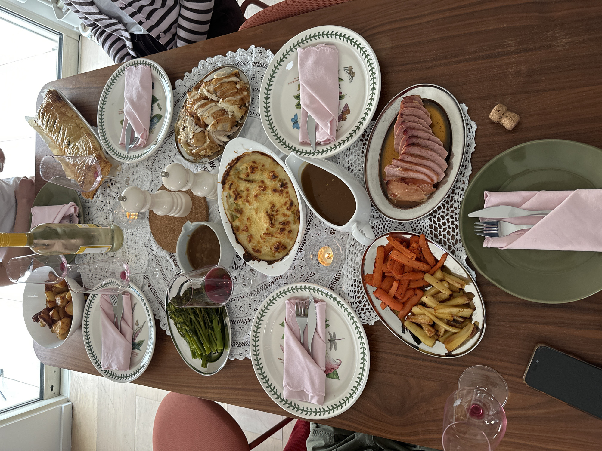 The image depicts a dining table laden with food, possibly for a festive meal. The table, made of wood, is partially covered with a white lace tablecloth. Several plates are arranged around the table, each with a light pink napkin and silverware. The plates are decorated with botanical patterns.
The food items include a loaf of bread, sliced chicken, a baked dish with a golden crust, a gravy boat with gravy, sliced ham, carrots and parsnips, roasted potatoes, and cooked green vegetables. Salt and pepper shakers are placed near the center of the table, along with a small candle. Several wine glasses, some filled with red wine, and a bottle of white wine are also present. A cork and a phone are on the table as well.
Two people are partially visible at opposite ends of the table, but they are not the focus of the image. One person is wearing a striped top. The location is likely a private residence. There is no information to identify the image as being in London, United Kingdom.