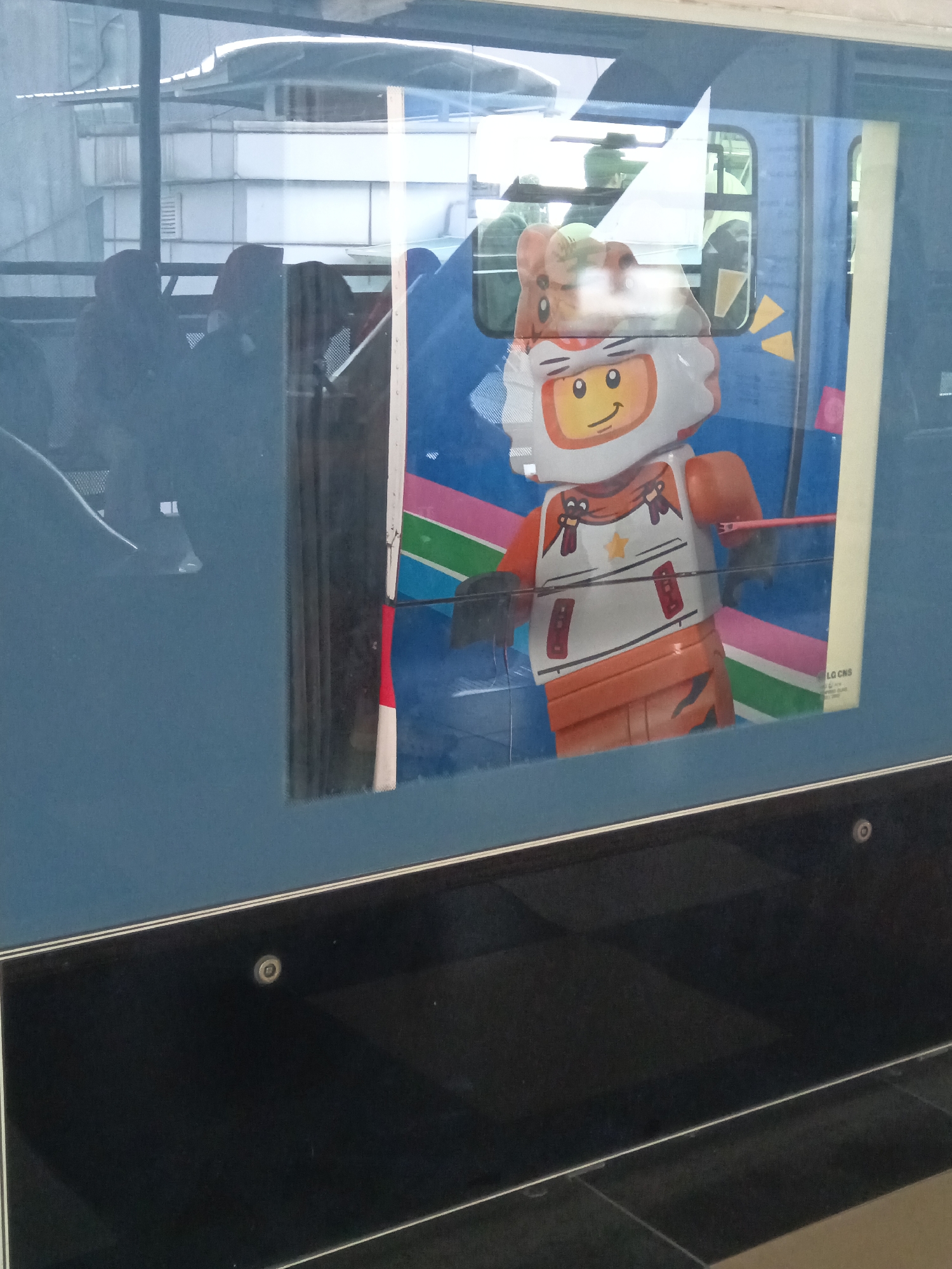 The image, captured at the KL Sentral LRT Station in Kuala Lumpur, Malaysia, shows a reflection in a glass window. The reflection reveals a blue train carriage with a large graphic decal of a Lego character. The character is a tiger-themed astronaut, wearing an orange and white suit with orange gloves and boots. The Lego character's helmet is shaped like a tiger's head with ears. Behind the Lego character, the interior of the train is visible, showing passengers with their backs to the camera. Some of these passengers are wearing headscarves. The exterior of the station, including its roof and structure, is also reflected in the glass. The foreground of the image is dominated by a dark, reflective surface, likely part of the station's architecture or the train window itself, with a visible circular fixture.