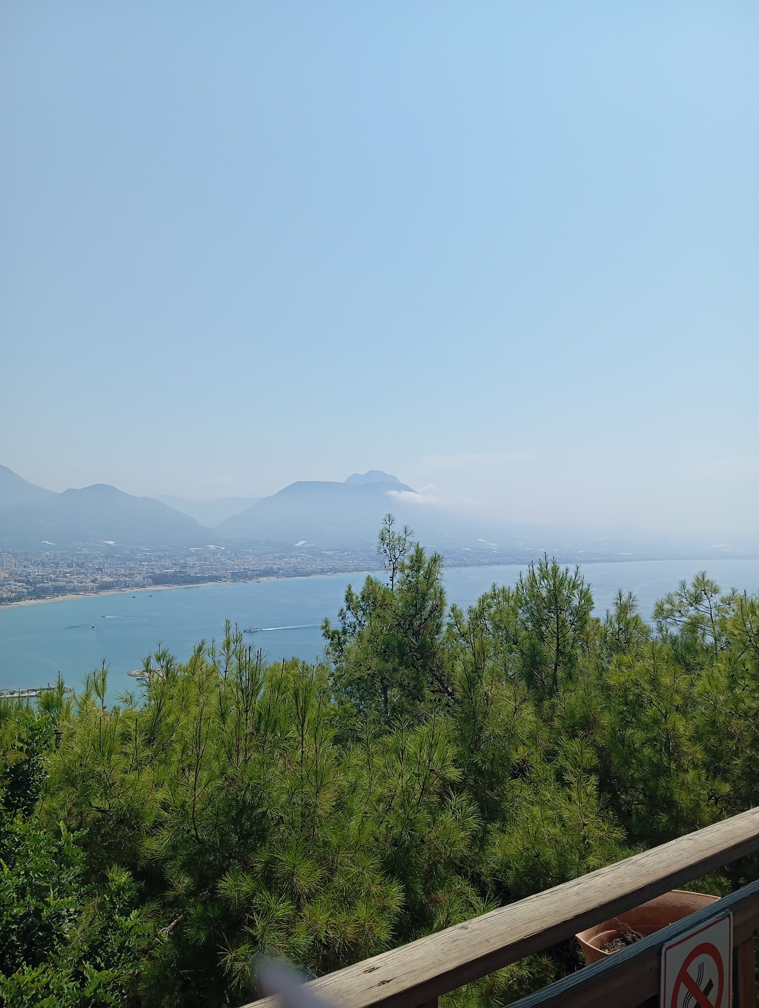An elevated, eye-level, wide outdoor shot captures a hazy, bright, coastal city nestled between mountains and the sea. In the foreground, a wooden railing runs diagonally from the bottom right corner upwards. To the left of the railing are lush, green pine trees, some of which obscure parts of the city in the mid-ground. The city itself features a dense collection of buildings, with a strip of beach and the blue oceanfront separating it from the distant, hazy mountains. Several boats are visible on the water. The sky above is a clear, light blue. A small, brown planter pot with dirt is situated on the railing in the lower right quadrant, and a red and white "no smoking" sign is attached to the railing.