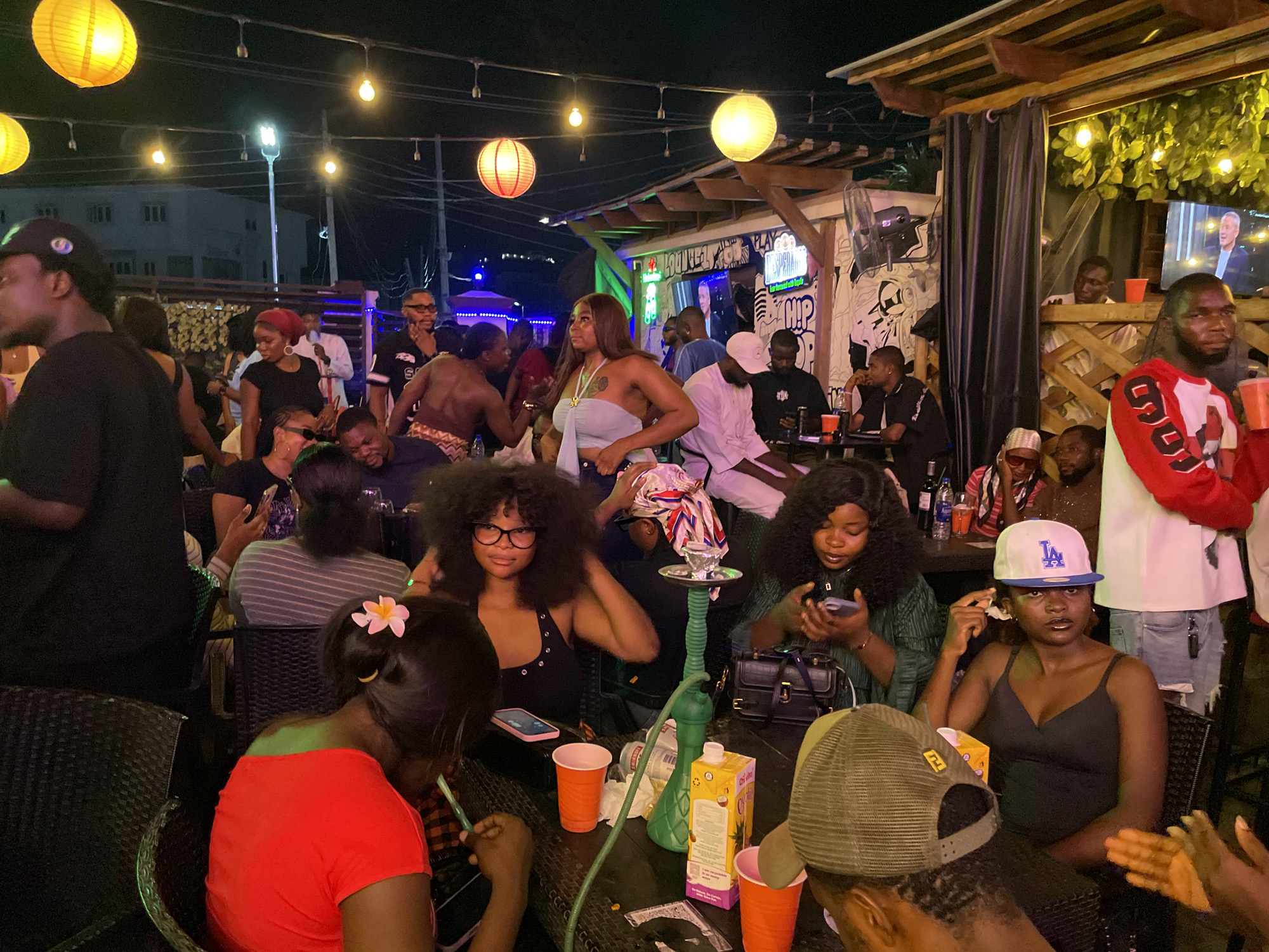 The image depicts a lively outdoor gathering, likely a social event or party, occurring at night. The location appears to be a bar or lounge, possibly in Lokoja, Nigeria, although this is not definitively confirmed in the image. The setting is illuminated by a string of overhead lights and lanterns.
Several people are visible, engaged in conversation and socializing. Some are seated around tables, while others are standing and interacting. Several people appear to be using their phones.
In the foreground, a group is gathered around a table with drinks, including juice. There is a hookah on the table. A woman in glasses with an afro is among the people at the table. A woman in a red shirt has a flower in her hair.
In the background, more people are visible. A television screen shows a man in a suit. The walls are decorated with graffiti.