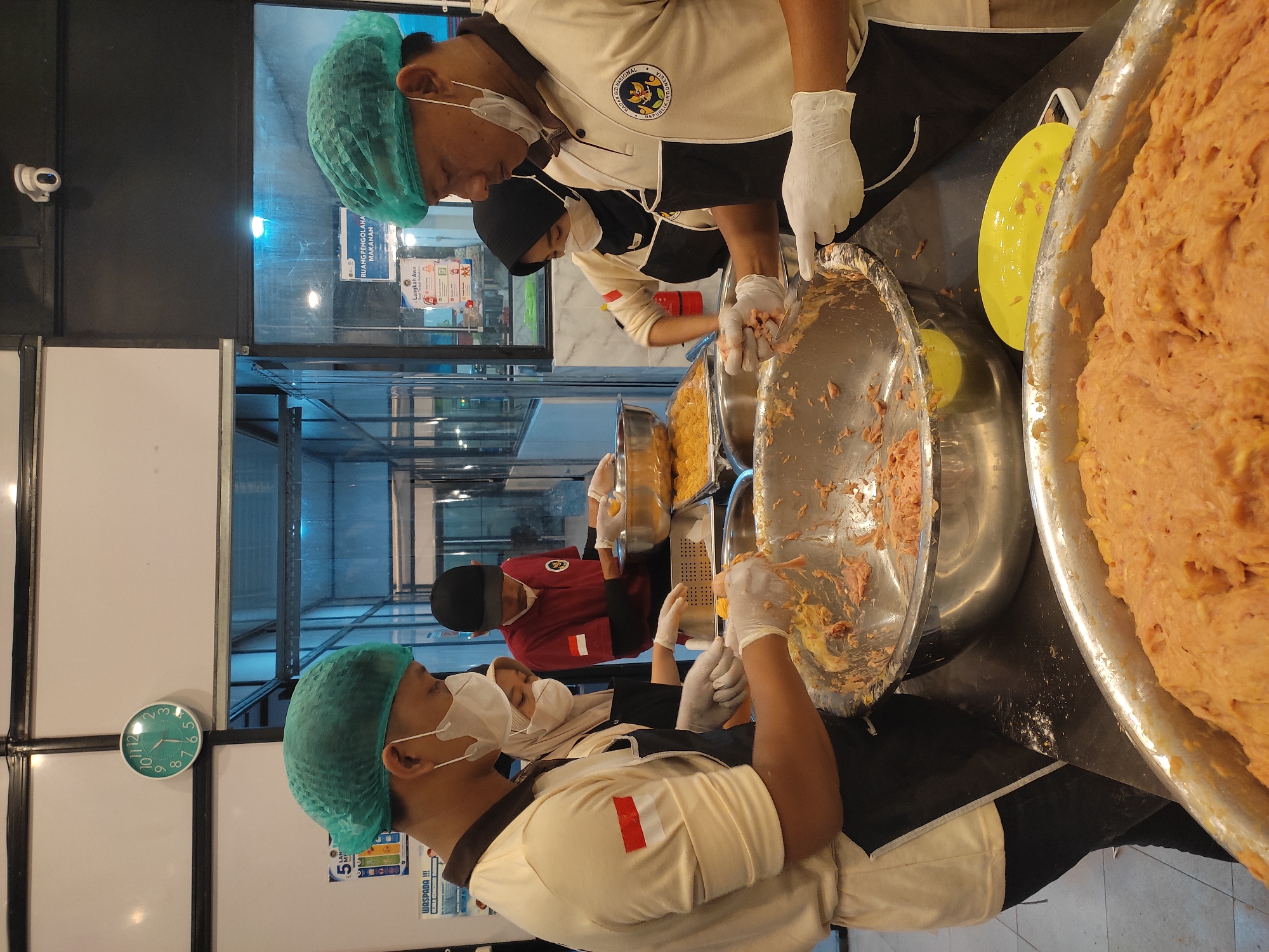 Four individuals, three women and one man, wearing green hairnets and white gloves, are engaged in food preparation. The man in the foreground, wearing a cream-colored polo shirt with an Indonesian flag patch, is actively mixing a pinkish mixture in a large stainless steel bowl. Another man, wearing a red shirt, stands behind him, also working with food. Two women are positioned behind the man in the foreground. One woman, wearing a headscarf and a cream-colored top with a black apron, is also working with food. The second woman, wearing a black headscarf and a white top with a black apron, is visible behind the first woman. The scene appears to be a commercial kitchen with stainless steel countertops and large metal bowls filled with food ingredients. A clock on the wall indicates it is 9:17. A security camera is visible mounted on the ceiling. Signs are posted on the wall, one of which reads "RUANG PENGOLAHAN MAKANAN" (Food Processing Room). The location is the BNN (Badan Narkotika Nasional) office in Ponorogo, Indonesia, as indicated by the logo on the man's shirt and the overall context of the BNN's activities.