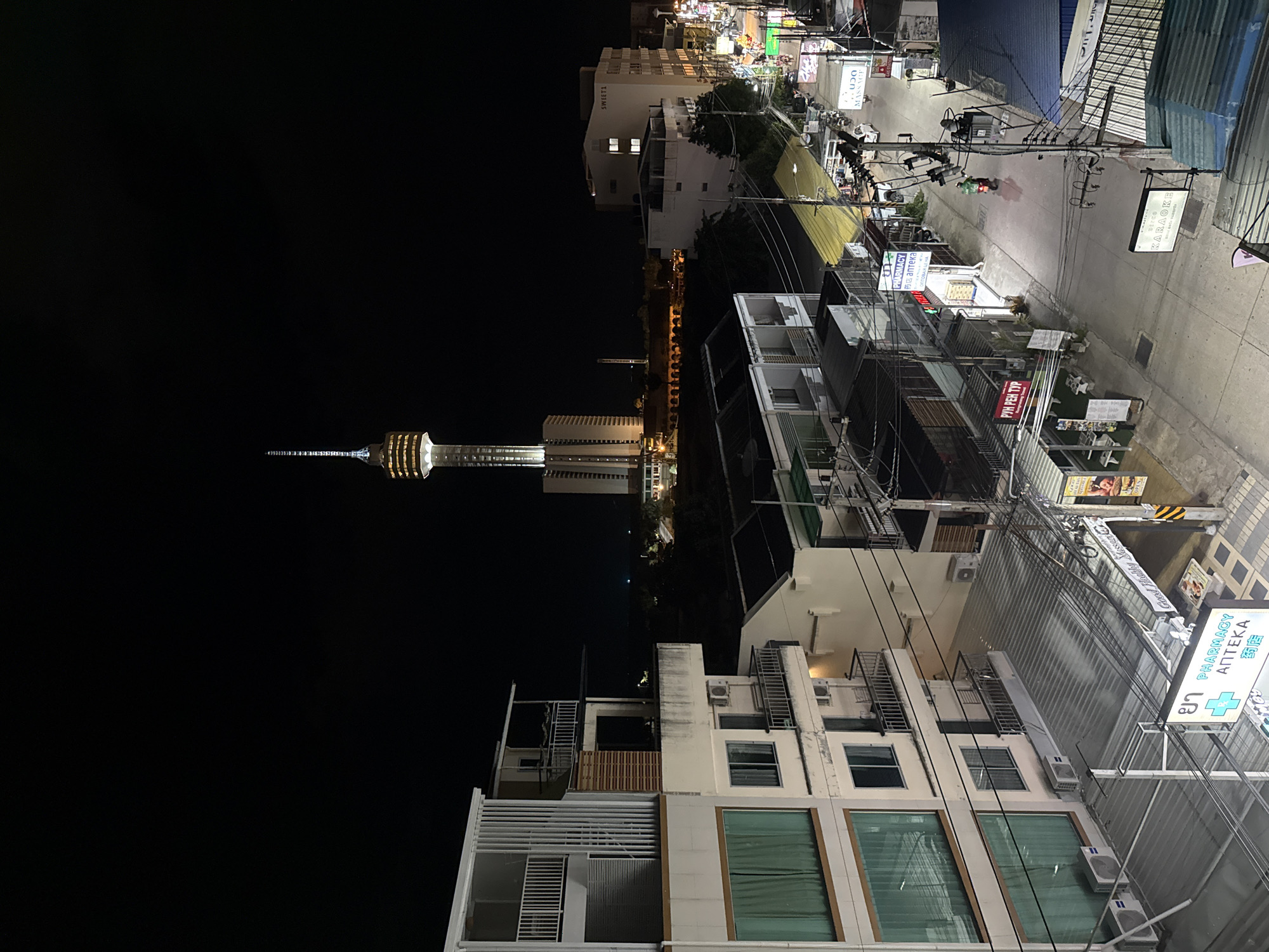 This media file captures a nighttime scene in Pattaya City, Thailand. The image is oriented vertically, showing a street scene with buildings and a prominent tower in the distance.
The street is lined with various establishments, indicated by numerous signs. Visible text includes "PHARMACY," "KARAOKE," and "MASSAGE" in multiple languages, suggesting a commercial and entertainment district. There are also signs with Thai script and Cyrillic text. Electrical wires crisscross overhead, a common sight in urban Asian settings. A motorcycle with a rider is visible on the street, indicating some level of activity.
In the background, a tall tower with illuminated sections stands against the dark night sky. The surrounding buildings are illuminated, casting a warm glow on the street below. The overall impression is of a bustling urban environment at night. The lighting suggests it is nighttime. The weather appears clear, with no visible rain or fog. The presence of a pharmacy sign with "ΑΠΤΕΚΑ" suggests a mix of local and tourist clientele.