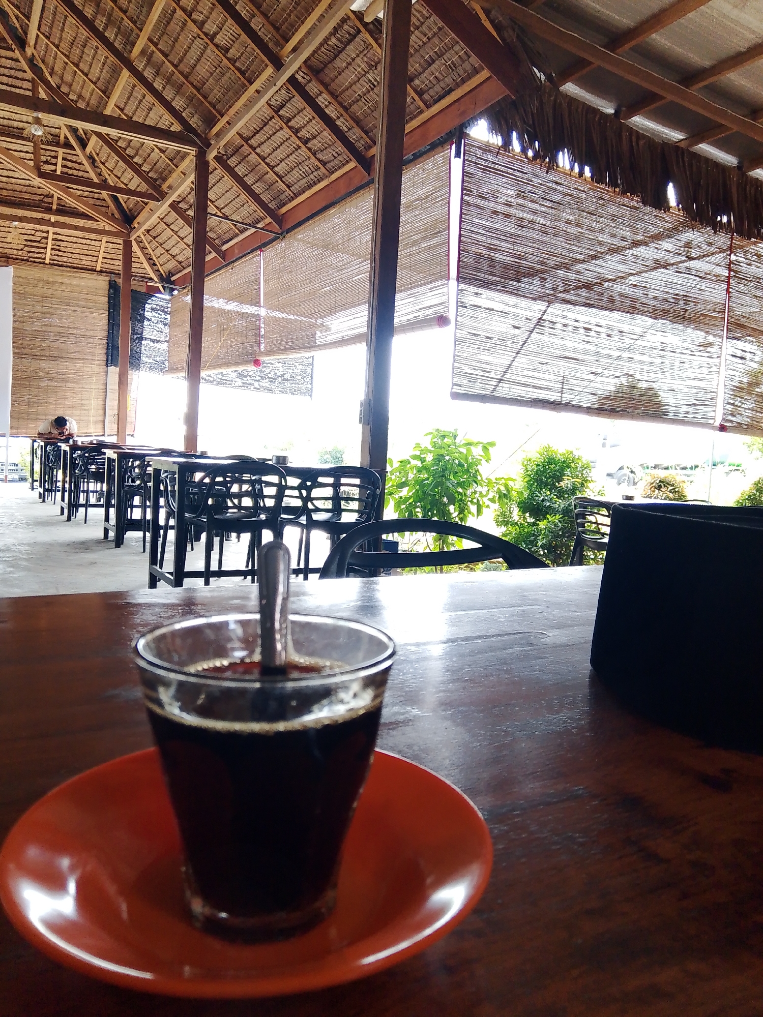 The image captures an outdoor seating area at a cafe in Meunasahmee, Indonesia, featuring a rustic, thatched roof and bamboo blinds. In the foreground, a glass of dark coffee sits on a wooden table, accompanied by a small orange saucer and a spoon. The background reveals a row of empty black tables and chairs, with a single person seated at one of the tables further down. Lush green plants are visible beyond the seating area, suggesting a tropical or subtropical environment. The lighting indicates it is daytime, with natural light filtering through the bamboo blinds.