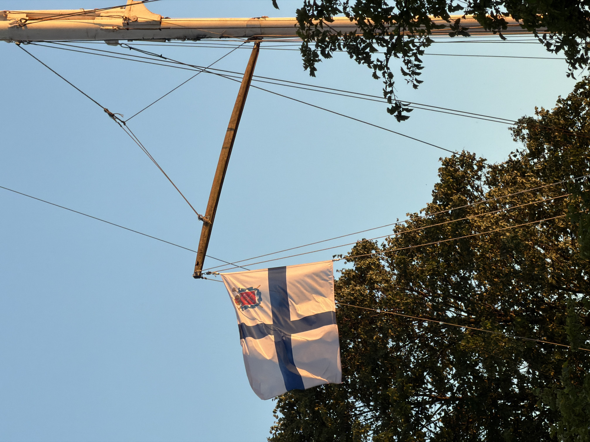 A white flag with a blue cross and a coat of arms in the upper left corner is attached to a flagpole. The flag is partially obscured by trees. The sky is clear and blue. The image appears to have been taken during the day, possibly in the late afternoon, given the warm light on the flagpole. The scene is likely located in Saint Petersburg, Russia, based on the flag's design, which resembles the city's flag.