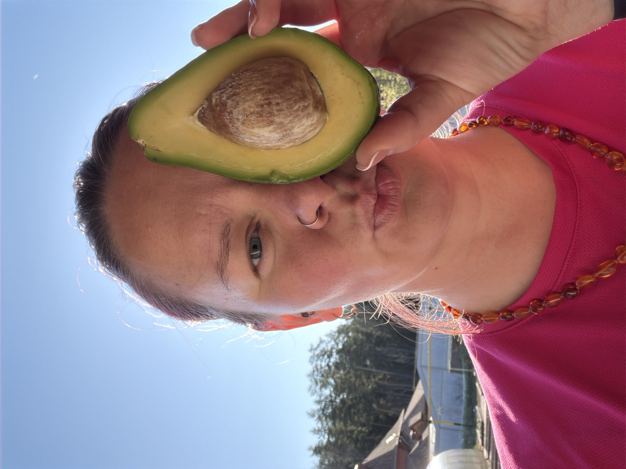 A woman in a pink shirt is holding half an avocado in front of her face, over her eye. She has a nose ring and a necklace of amber-colored beads. The background shows a blue sky, trees, and a building with a greenhouse. The photograph may have been taken outdoors. The city and country of Chaikovsky, Russia are not readily apparent in this image, and cannot be determined.