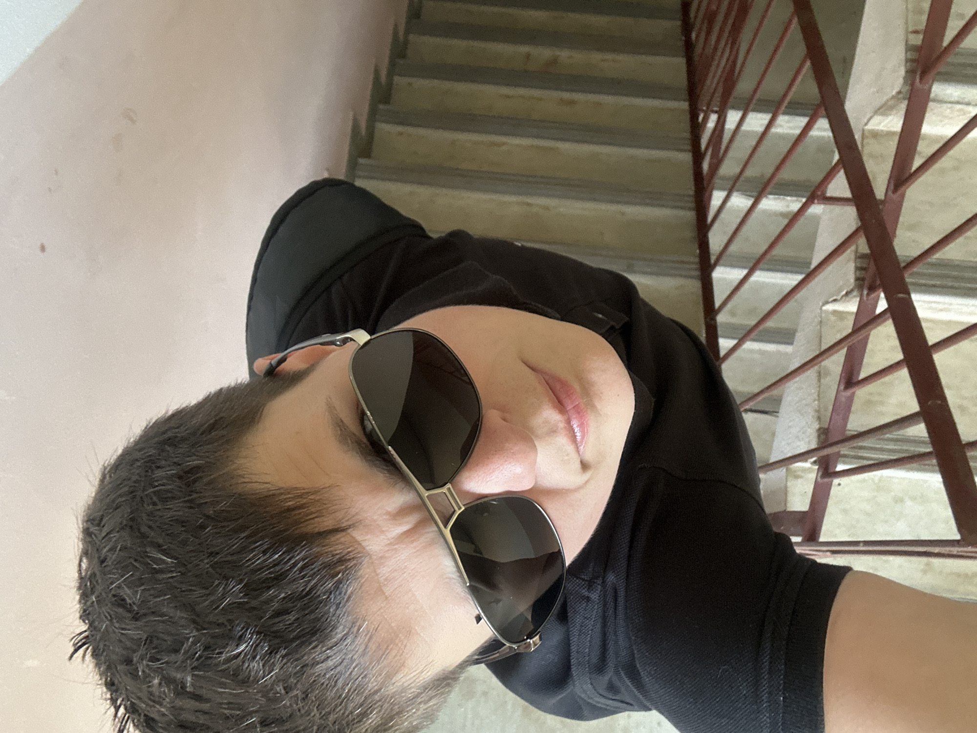 A person is captured from a high angle, looking up towards the camera, on a concrete staircase with a red metal railing. The person has short, dark, messy hair and is wearing dark sunglasses and a black polo shirt. Their face is slightly wrinkled in a way that suggests a squint or a frown. The staircase appears to be outdoors, as indicated by the lighting and the texture of the concrete. The scene is devoid of any distinguishing landmarks or signage that would allow for precise location identification within Uzhhorod, Ukraine, or any other city. The overall environment is functional and urban, suggesting a utilitarian architectural setting rather than a tourist attraction or a specific neighborhood.