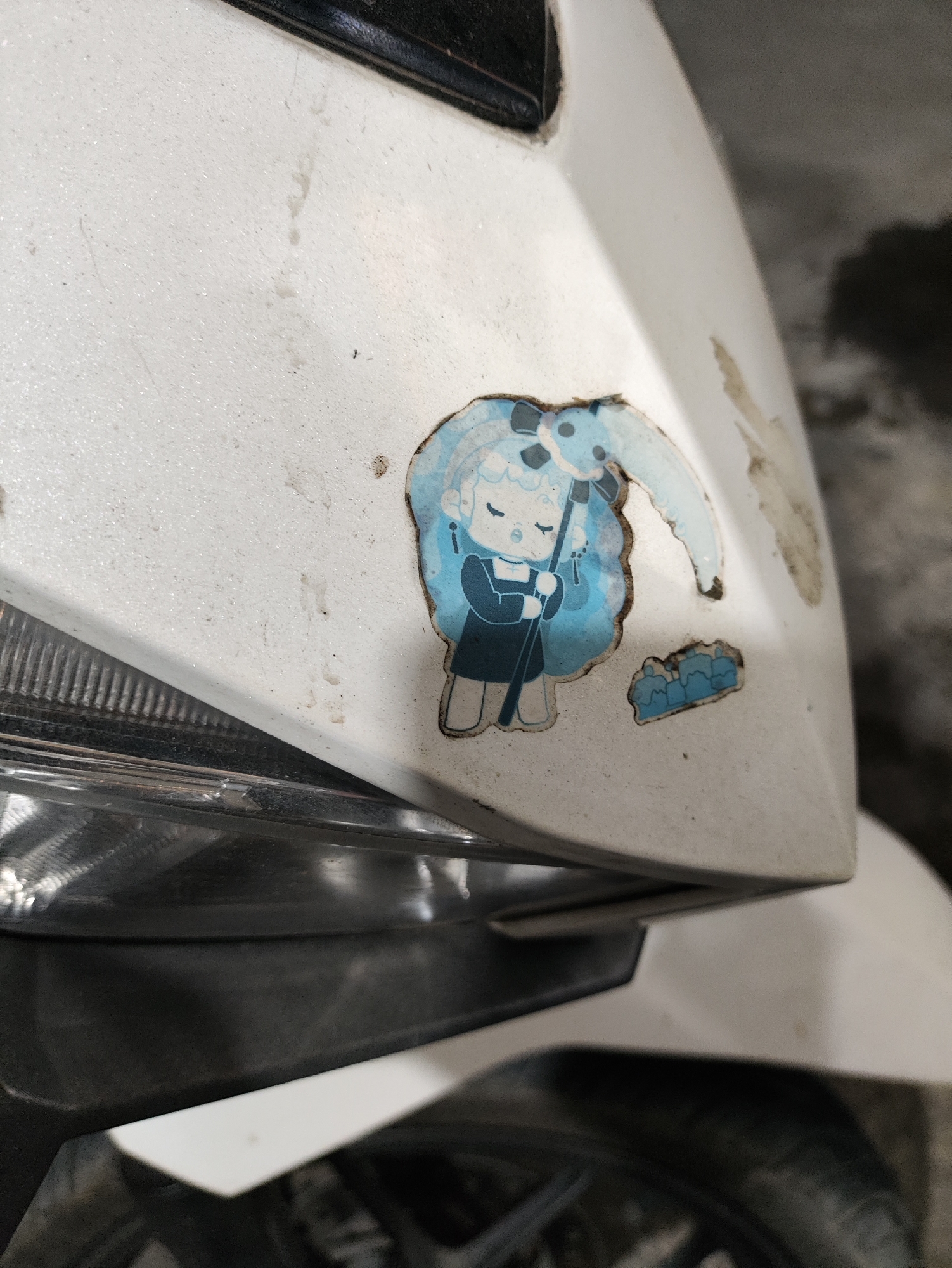 A sticker of a cartoon character is attached to the front white panel of a scooter. The character appears to be a young girl with blue hair in a bun, wearing a blue dress and holding a dark stick. There is also a smaller blue sticker below the character depicting a cluster of rounded shapes. The scooter is dirty, with dust and grime visible on the white surface. The lower left of the image shows the edge of a black headlight. The background is dark and out of focus, suggesting an indoor or dimly lit environment. The scene is located in Kepung, Indonesia.