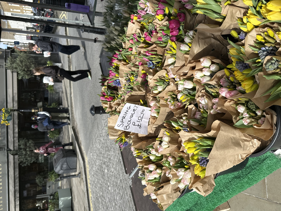The image depicts a vibrant outdoor flower stall situated on a cobbled street in Chelsea, London, United Kingdom, under bright daylight. In the immediate foreground, numerous "Spring Bouquet" arrangements, primarily consisting of tulips and hyacinths in shades of yellow, pink, white, and blue/purple, are individually wrapped in brown paper and displayed in dark containers on a green-surfaced stall. A handwritten white sign states "Spring Bouquet £10.00."
In the midground, a cobbled street is visible with pedestrian traffic. A woman with dark hair, wearing a dark top and black flared trousers, walks left, carrying a white object. Behind her, a man with dark hair, dressed in a dark hoodie and dark trousers, also walks left while eating from a white object. Further back on the pavement, three individuals stand near a building; one wears a burgundy jacket, another a dark top with a blue bag, and a third a light jacket. A black bollard is present in the street.
The background features classic London architecture, including a building with dark awnings and the distinct sign "CHELSEA TEXTILES" visible above. Other shopfronts, lampposts, and parked vehicles line the street further down the perspective, indicative of an urban commercial area.