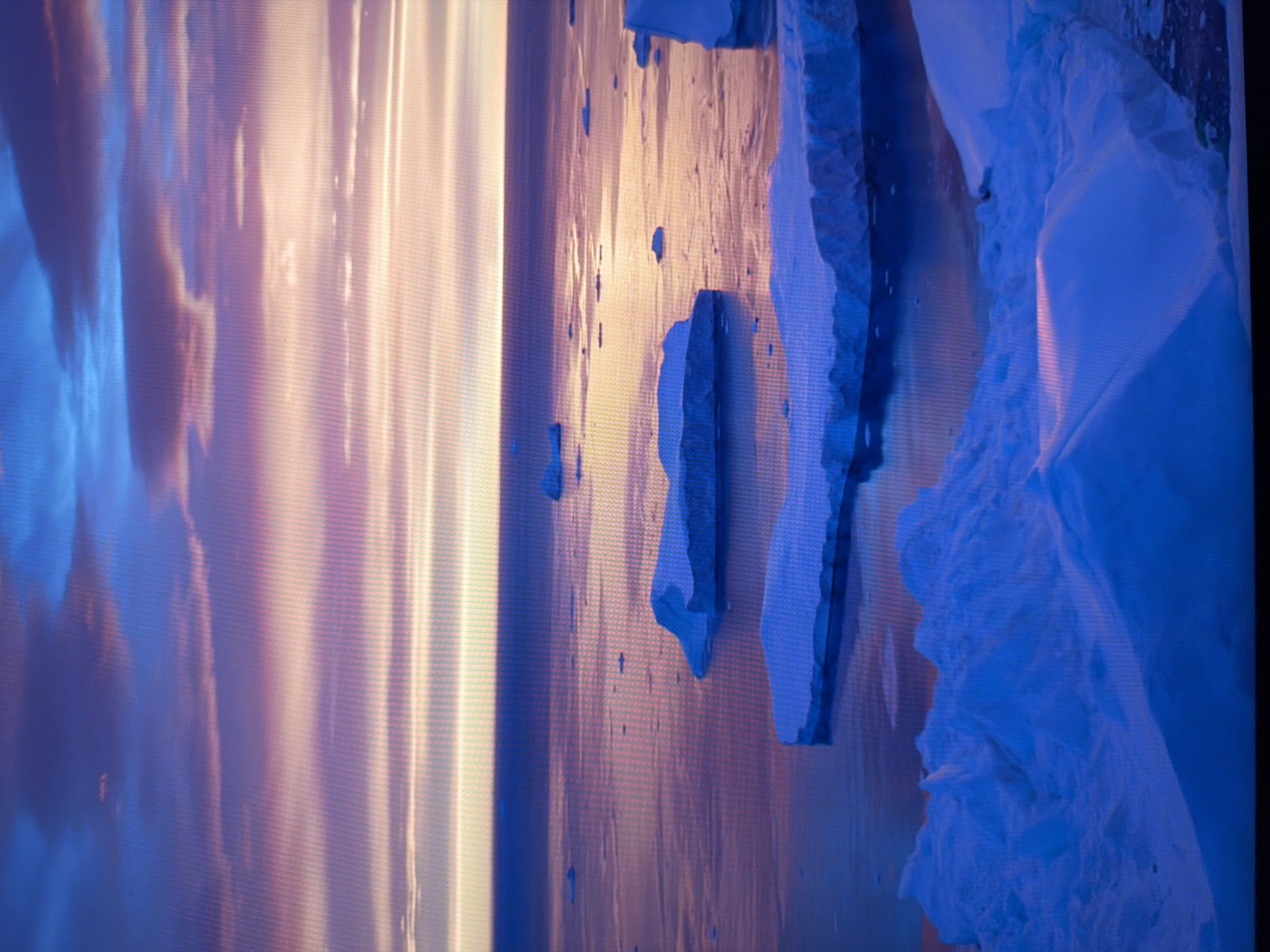 This is a photograph of a serene icy landscape at dusk. The sky is a gradient of deep blues and purples, streaked with lighter pinks and oranges, suggesting the sun is low on the horizon. Below the sky, a vast expanse of calm water reflects the colors of the sky, creating a soft, ethereal glow. Scattered across the water are numerous icebergs of varying sizes, their white and blue surfaces illuminated by the fading light. Some are small, solitary pieces, while others are larger, more substantial formations. In the foreground, a massive iceberg dominates the view, its textured, snow-covered surface a brilliant white. The overall scene conveys a sense of peace and immensity, characteristic of an Arctic or Antarctic environment. There are no people or man-made structures visible in this image. The photograph does not depict any location in the city of Kyiv, Ukraine.