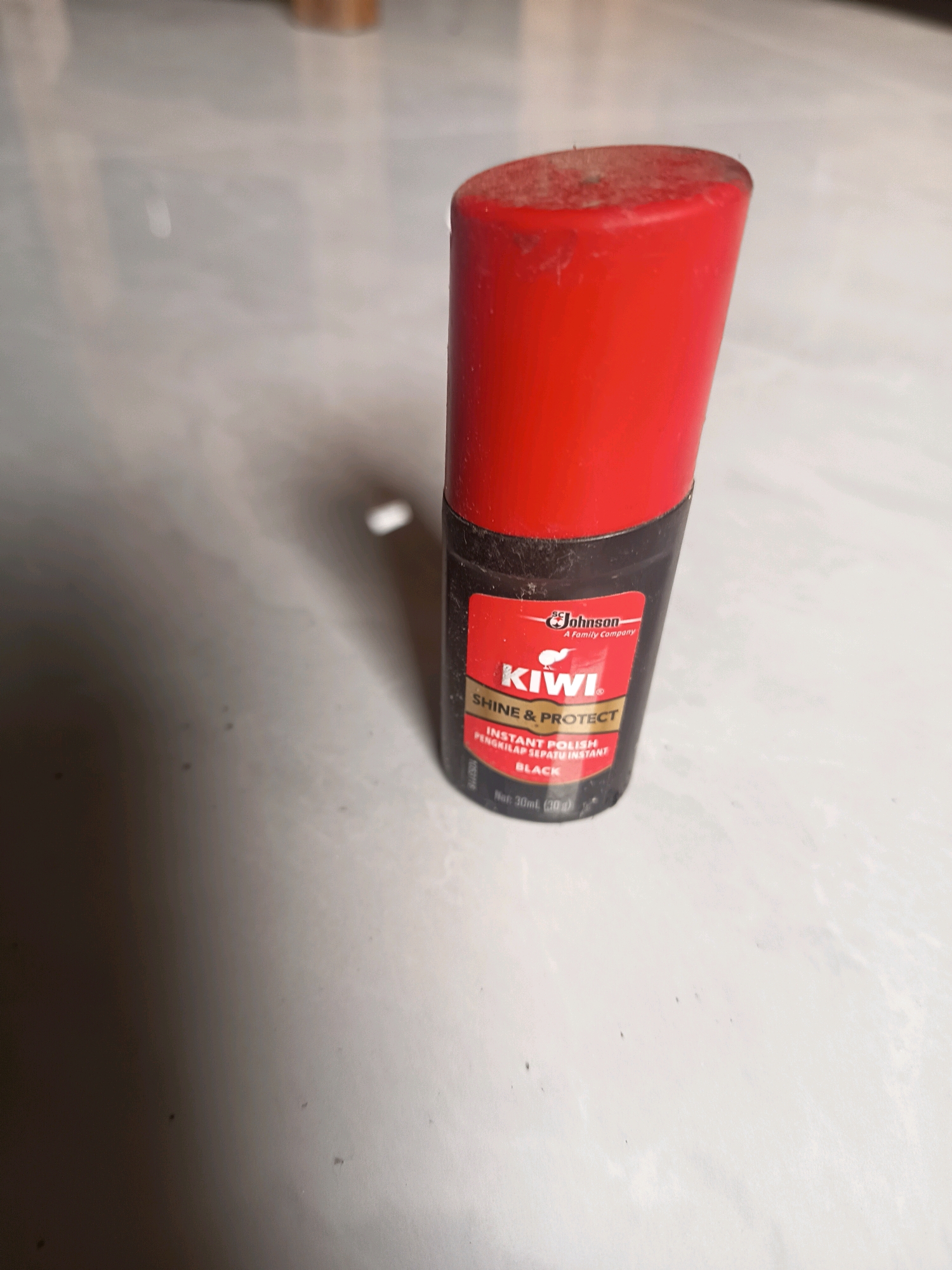 An off-center, slightly high-angle shot shows a bottle of Kiwi Instant Polish in Black. The bottle has a red cap and a black body with a red label. The label reads "Johnson A Family Company," "Kiwi," "SHINE & PROTECT," "INSTANT POLISH," "PENGKILAP SEPATU INSTANT," and "BLACK." Below the word "BLACK" it says "Net: 30mL (30 g)." The bottle is standing upright on a light-colored, speckled surface. The lighting creates a dark shadow on the left side of the image. There is a faint, out-of-focus background that appears to be a blurred surface. The presence of this product suggests a location where shoe care might be relevant, such as a household, shop, or area where people maintain their footwear. This scene is not specifically identifiable as being in Sidorejo, Indonesia, based on visual cues alone.