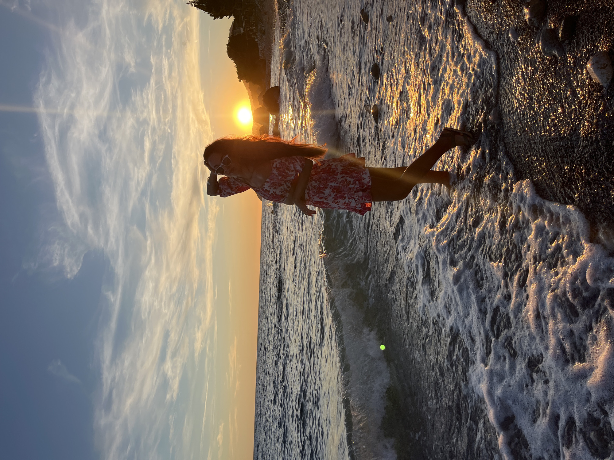 The image depicts a young woman standing on a pebble beach in Bar, Montenegro, at sunset. She is wearing a short, floral-patterned dress and white sunglasses, with her right arm raised and hand touching her hair. The sun is low on the horizon, casting a warm, golden glow across the scene and reflecting on the water. Gentle waves are breaking on the shore, creating a foamy edge. In the background, a silhouette of trees and structures can be seen on a distant hillside. The sky is a blend of blue and orange hues, with wispy clouds illuminated by the setting sun. The tide is low, revealing wet pebbles and stones on the beach.