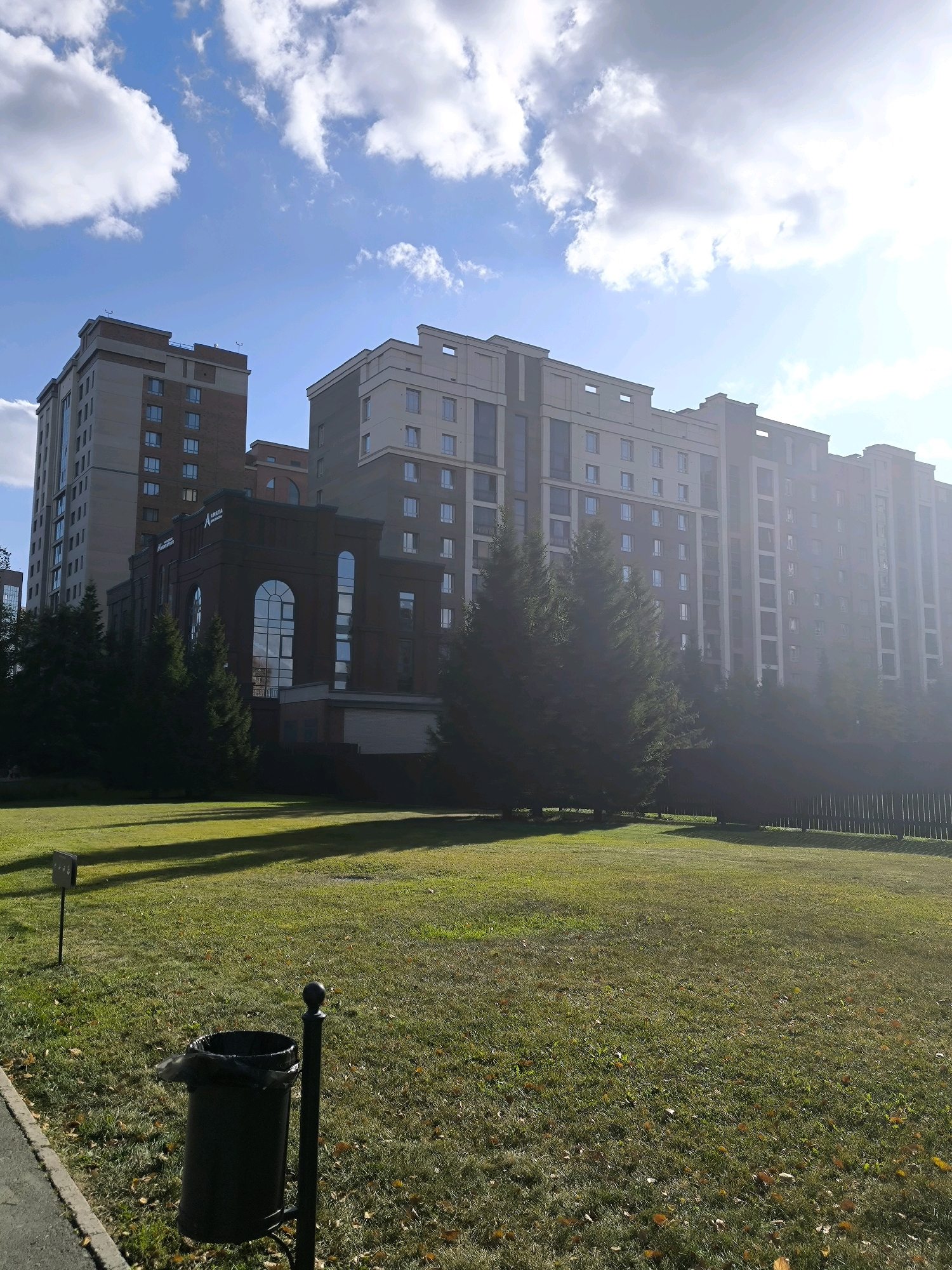 The image depicts a view of modern apartment buildings and a smaller brick building with arched windows. The scene is set in a city park with green grass, scattered fallen leaves, and several evergreen trees. A black trash can on a stand is visible in the foreground, as is a small signpost. The sky is blue with scattered white clouds, and the sun is shining brightly, creating shadows on the grass. The architecture suggests a contemporary urban development. This image was taken in Novosibirsk, Russia.