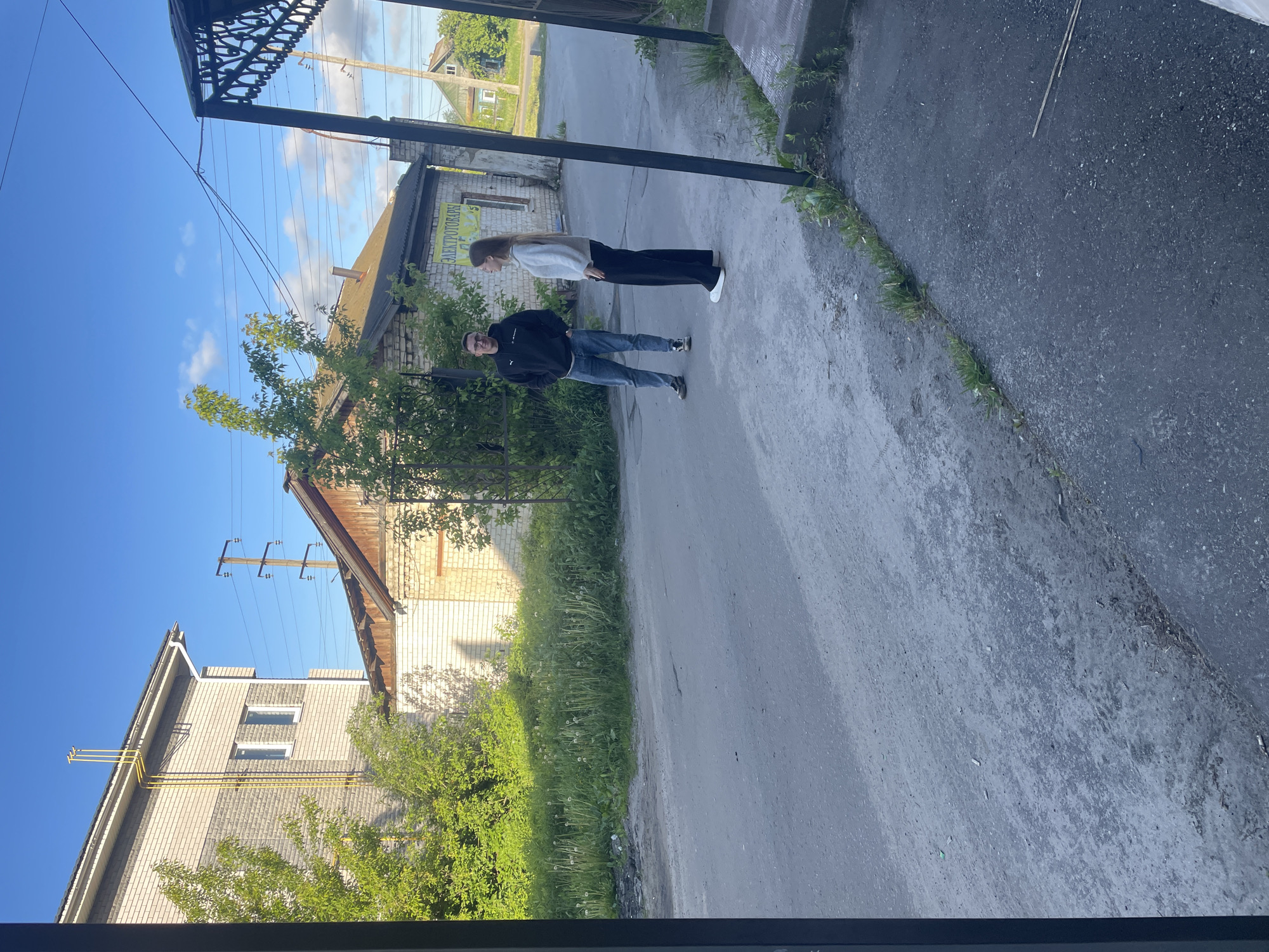 The image presents an outdoor scene on a clear, bright day, likely morning or afternoon, in Semyonov, Russia. Two individuals stand on an asphalt path. A man, closer to the viewer, wears a dark hooded jacket, blue jeans, and glasses, looking towards the camera. Behind him, a woman with long hair is dressed in a light-colored long-sleeved top and dark pants, also facing the camera.
The setting includes buildings, green foliage, and utility infrastructure. On the left, a brick building with a pitched roof displays prominent yellow gas pipes along its facade. The path is bordered by lush green bushes and trees. On the right, another building, constructed from brick and wood, features a yellow sign with the Cyrillic text "ЭЛЕКТРОТОВАРЫ" (Elektrotovary), translating to "Electrical Goods." Power lines and utility poles are visible against the blue sky, which has scattered white clouds. A decorative dark metal structure, possibly an awning or gate, is partially visible in the upper right. The asphalt path shows some wear, with grass and weeds growing along its edges. The scene captures a moment on a quiet, local street with characteristic Russian architecture and infrastructure.