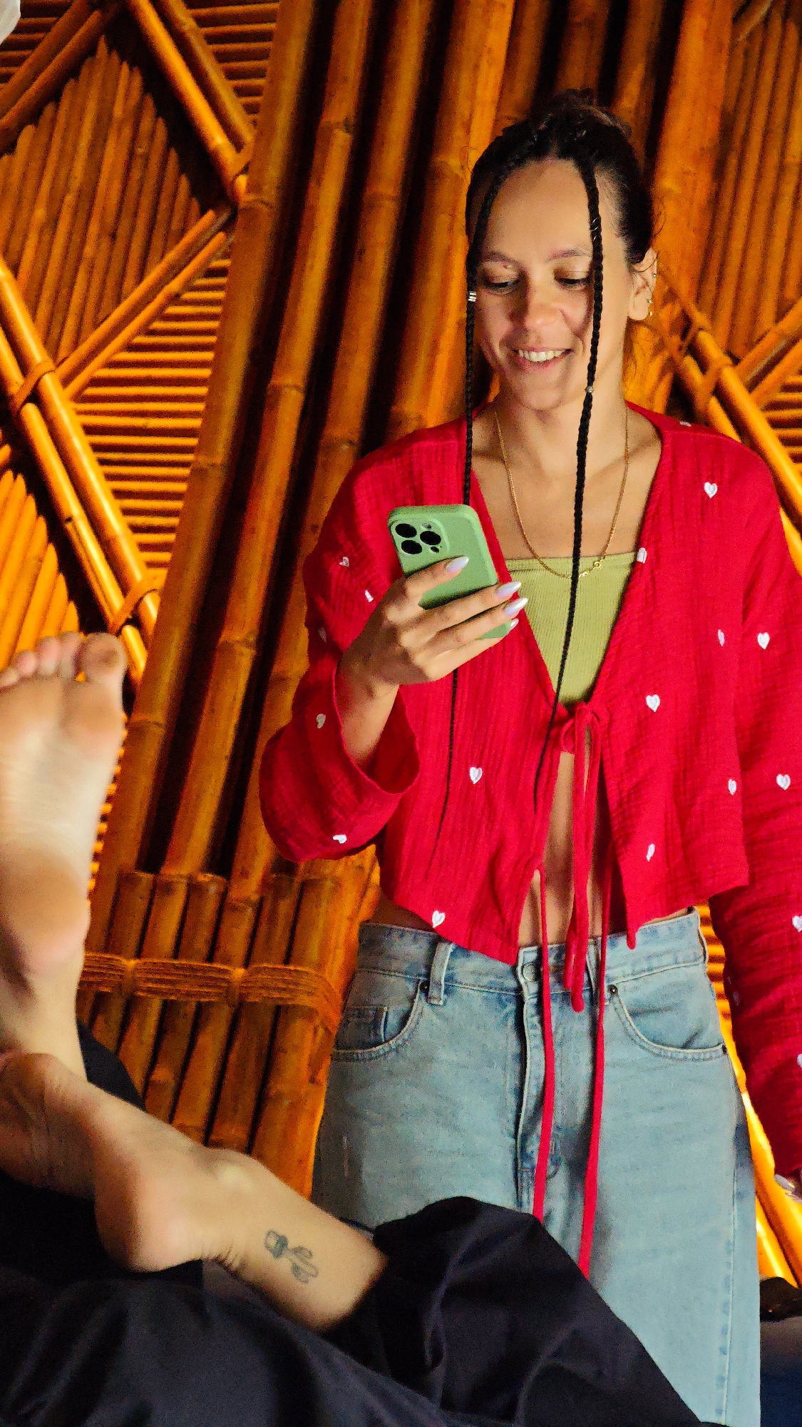 An adult female is depicted standing in a warm-toned, indoor environment, stated to be in the city of Mu Cang Chai, Vietnam. She has dark hair styled with two prominent braids framing her face and silver rings on the braids. She is smiling and looking down at a bright green smartphone, held with both hands. Her attire consists of a red, long-sleeved, cropped top adorned with small white heart patterns, tied at the front, revealing a light green ribbed tank top underneath. She is also wearing light blue denim jeans and a thin gold chain necklace. In the lower-left foreground, the bare foot and lower leg of a second individual are visible, resting on a dark fabric surface. A small, dark tattoo, appearing to be a cactus or plant, is present on the visible ankle of this second individual. The background features a structural wall constructed from light brown to orange-toned bamboo poles or wooden slats, arranged in vertical and diagonal patterns, illuminated by warm lighting.