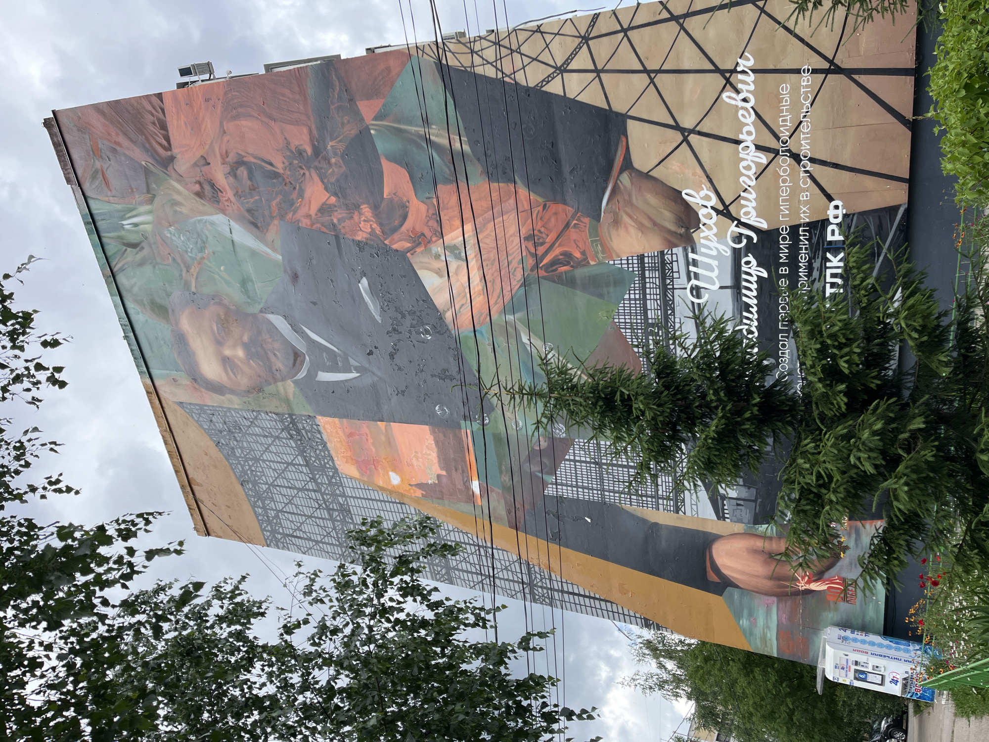 The image shows a large mural on the side of a building in Nizhny Novgorod, Russia. The mural depicts a portrait of a man in formal attire, with abstract colorful elements and architectural blueprints in the background. Text in Russian is visible on the mural, identifying the subject as "Shukhov Vladimir Grigoryevich" and stating that he created the world's first hyperboloid structures and applied them in construction. The building itself appears to be a multi-story residential or office building. Trees and foliage partially obscure the lower part of the mural and the building. A street, a small kiosk, and a fence are visible in the foreground. The sky is overcast.