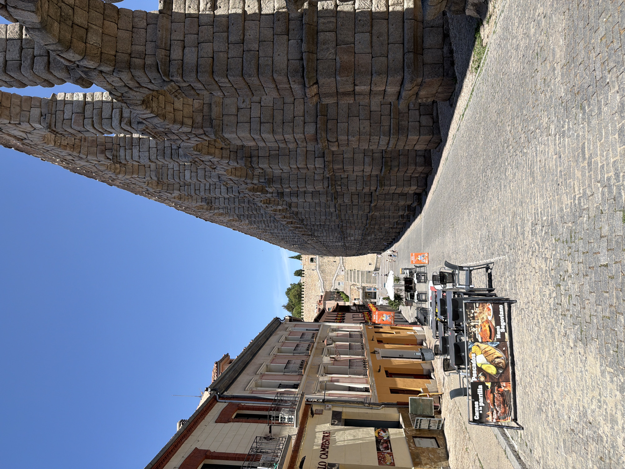 The image shows a street scene in Segovia, Spain, dominated by a section of the ancient Roman aqueduct. The aqueduct, constructed from large, precisely cut stone blocks, forms a towering arc on the right side of the frame, casting a shadow onto the cobblestone street below. To the left of the aqueduct, a row of buildings lines the street, featuring a mix of architectural styles. These buildings have facades with multiple stories, balconies with wrought-iron railings, and window shutters. Several outdoor tables and chairs are set up on the sidewalk in front of the buildings, suggesting a restaurant or cafe area. A black and silver A-frame sign with images of food, including grilled meats and fish, is prominently displayed in the foreground, advertising "Carnes a la parrilla" (grilled meats), "Pescados" (fish), and "Menú Segoviano" (Segovian Menu). Several people are walking in the distance along the street and towards a set of stairs leading up to a fortified wall. The sky is a clear, bright blue, indicating a sunny day. The street itself is paved with cobblestones, and a small strip of green grass is visible along the base of the aqueduct.