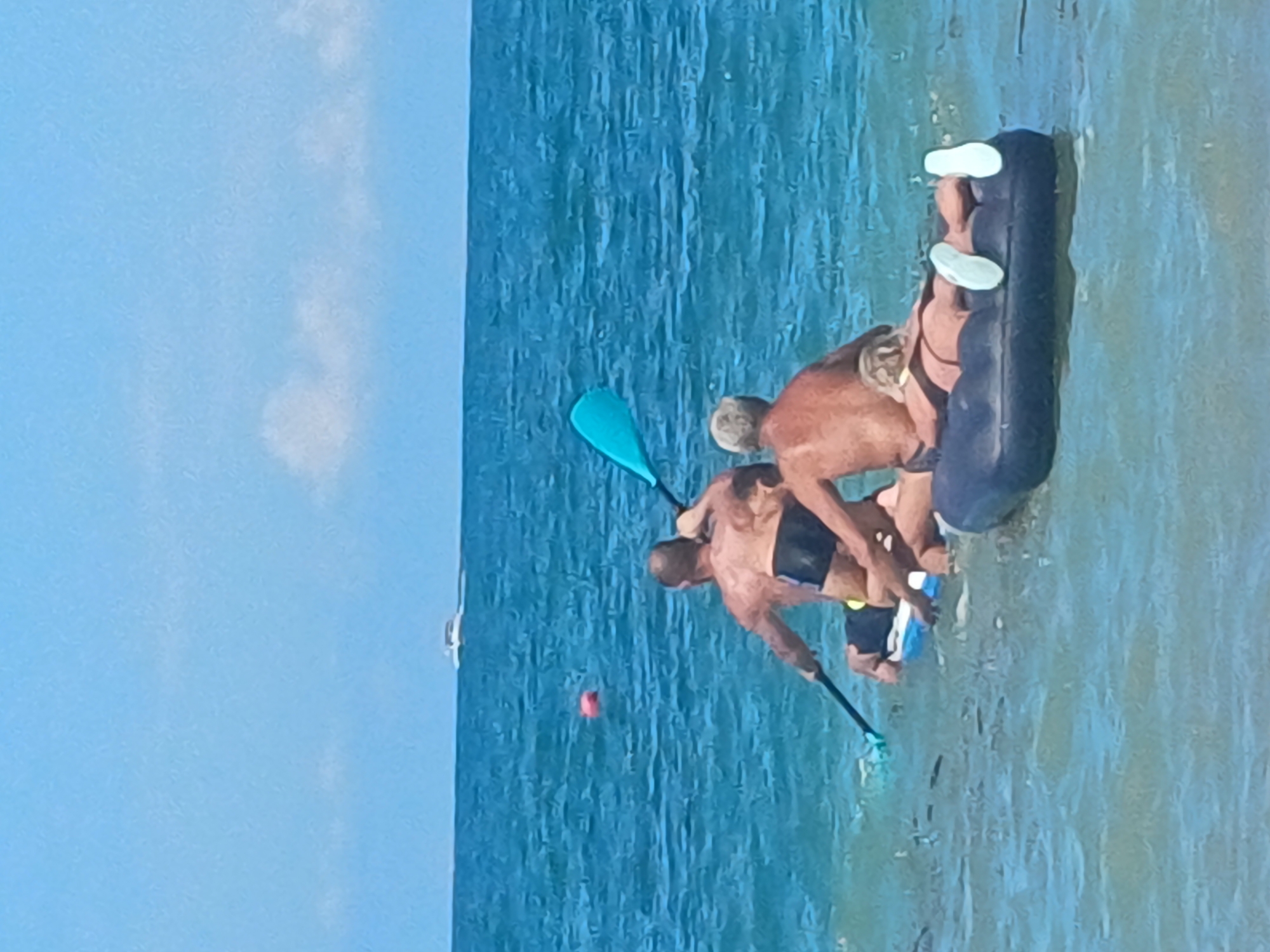 Two men are on a paddleboard in the water, one paddling with a turquoise paddle, the other appears to be sitting. In front of the paddleboard, an inflatable raft is floating, on which a person is lying down. The raft has white-soled shoes on it. The ocean is blue with some ripples, and the sky is clear and blue with some white clouds. A small boat is visible in the distance on the horizon. There is also a red buoy in the water to the left of the paddleboard. The scene appears to be a beach or coastal area, possibly in Sochi, Russia, given the blue water and clear sky, which are characteristic of the region.