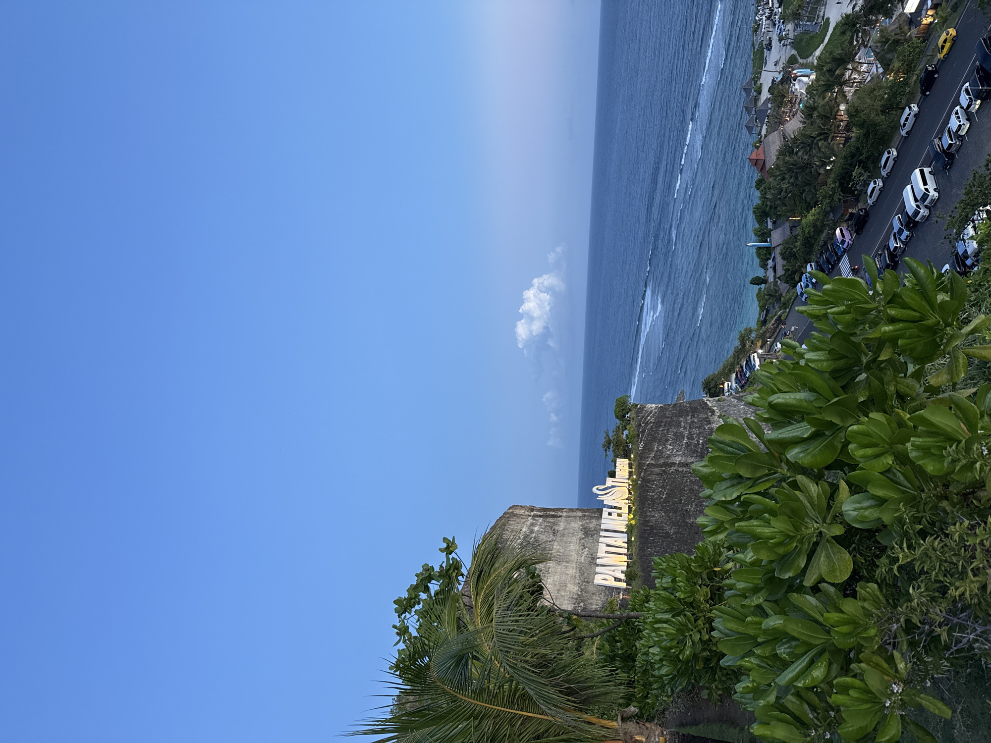 The photograph shows a view of Pantai Melasti, located in Ungasan, Indonesia. The scene includes the ocean, a cliffside with a sign that reads "PANTAI MELASTI", and various types of vegetation. Along the coastline, there is a road with vehicles parked alongside. The sky is a gradient of blue.