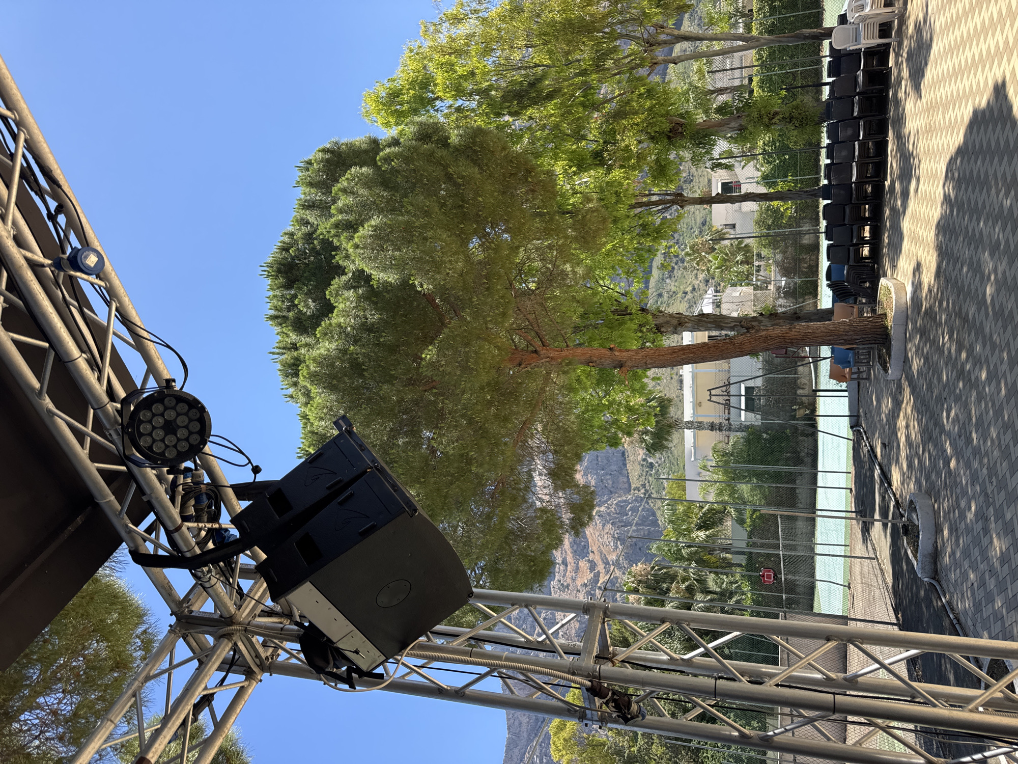 An outdoor event setup is depicted under a clear blue sky in Isola delle Femmine, Italy. In the foreground, a metal truss structure supports sound equipment, including large black speakers, and lighting fixtures. To the right, rows of empty chairs are arranged on a paved area, facing a tennis court beyond a fence. Lush green trees, including a prominent pine tree with a reddish trunk, dominate the midground. In the background, buildings and rugged hills are visible. The scene suggests preparation for an outdoor gathering or performance.
