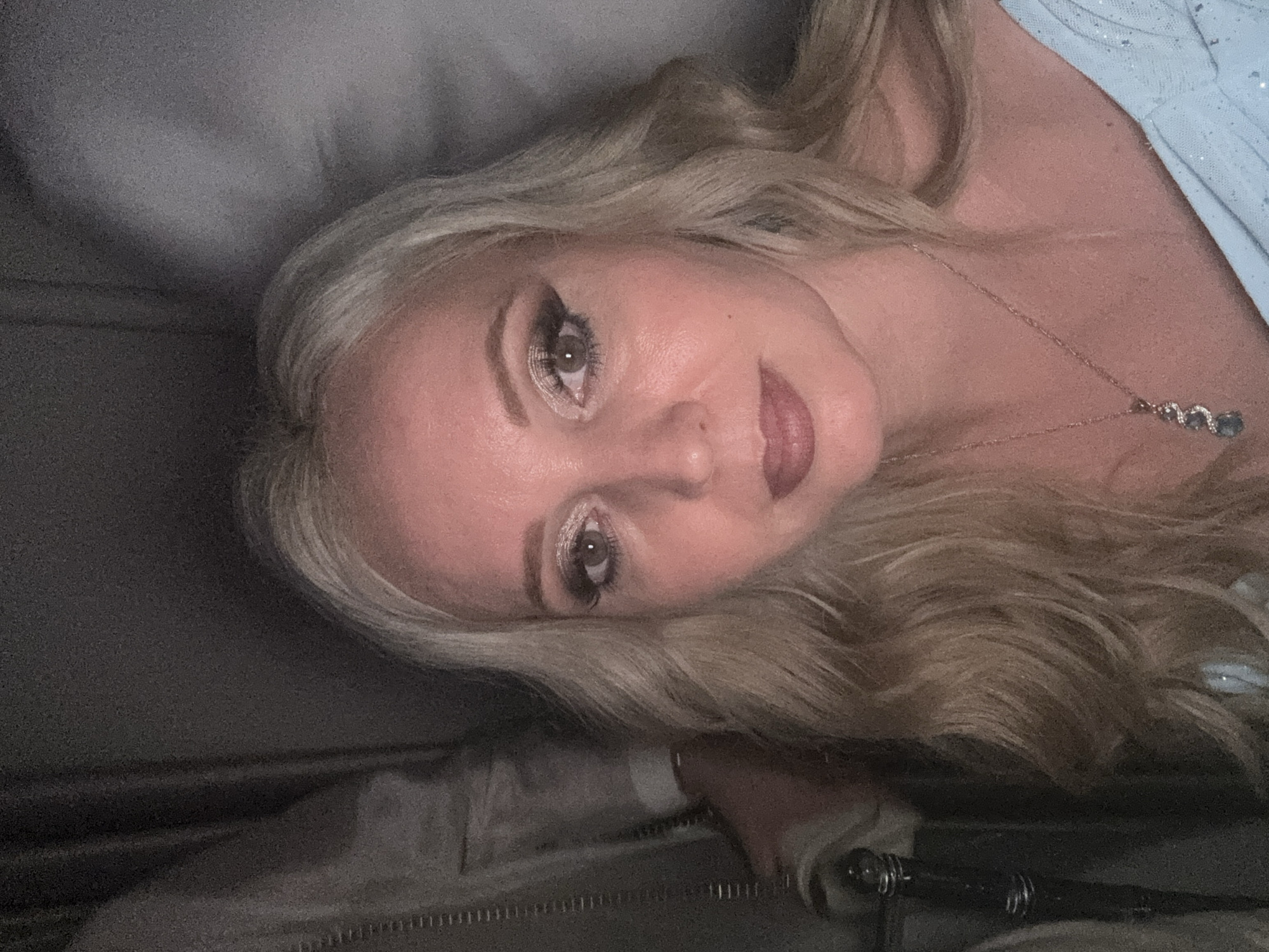 A close-up, waist-up selfie of a blonde woman with wavy hair, wearing a light blue dress and a necklace. She has on heavy makeup, including eyeshadow and eyeliner. She is indoors, against a dark background with some light filtering in from the right. The location in Komsomolsk-on-Amur, Russia, is unknown.