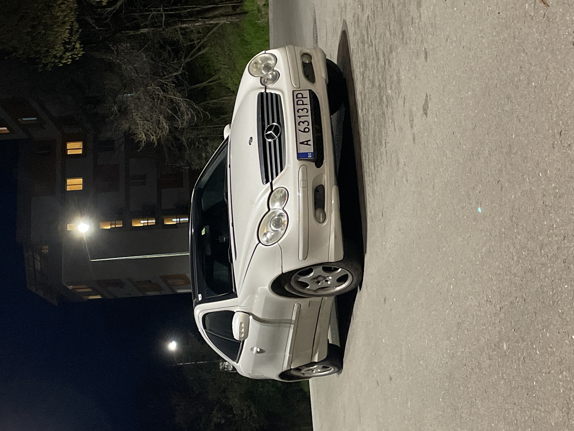 A white Mercedes-Benz C-Class is parked on an asphalt street in Veliko Tarnovo, Bulgaria, at night. The car is facing the camera, and its license plate reads "A 6313 PP." In the background, a building with illuminated windows can be seen. Trees and a streetlight are also visible.