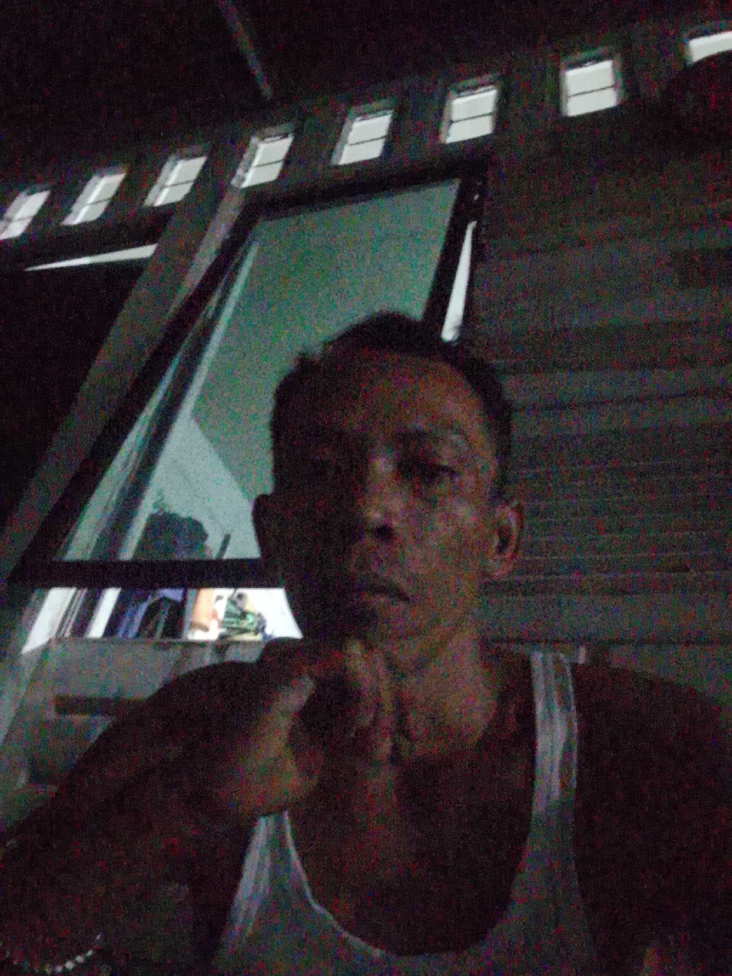 The image shows a person in a white tank top with their hand touching their chin. They are indoors, with a partially open window frame behind them. The window reflects light, possibly from outside. The background appears to be a wall made of wooden planks. The picture has a grainy, low-light quality. Based on the prompt's inclusion of Bukit Ketok, Indonesia, it can be inferred that the photo was taken in this location.